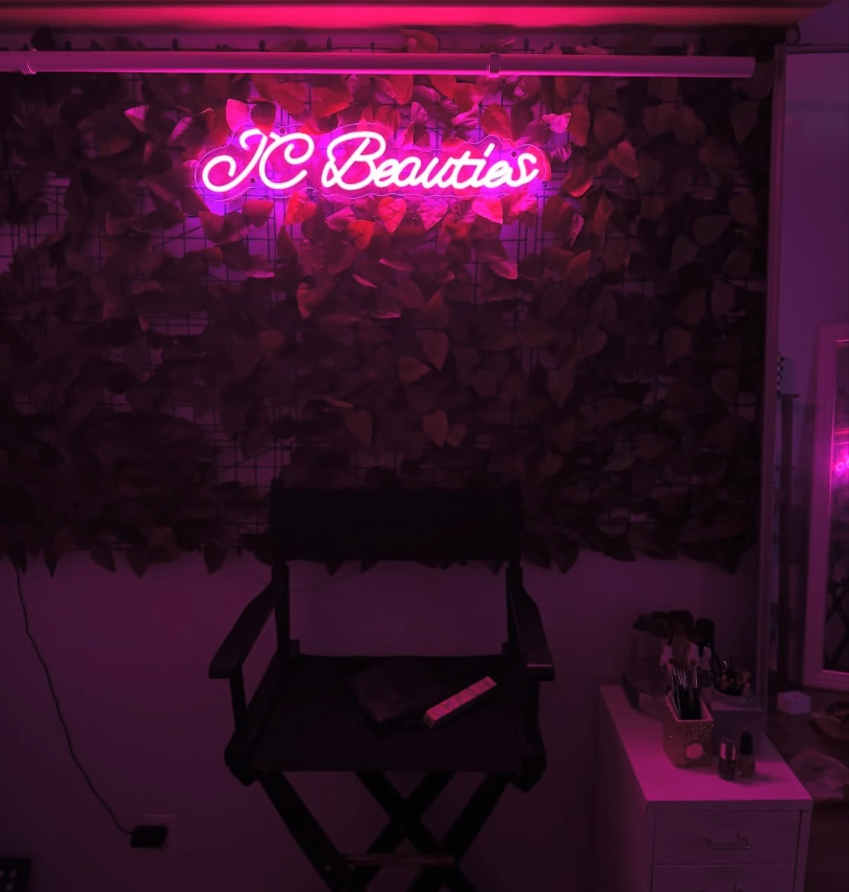 Neon pink sign reads 'JC Beauties' above a dark wall with plants, a black chair, and a small table with beauty products.