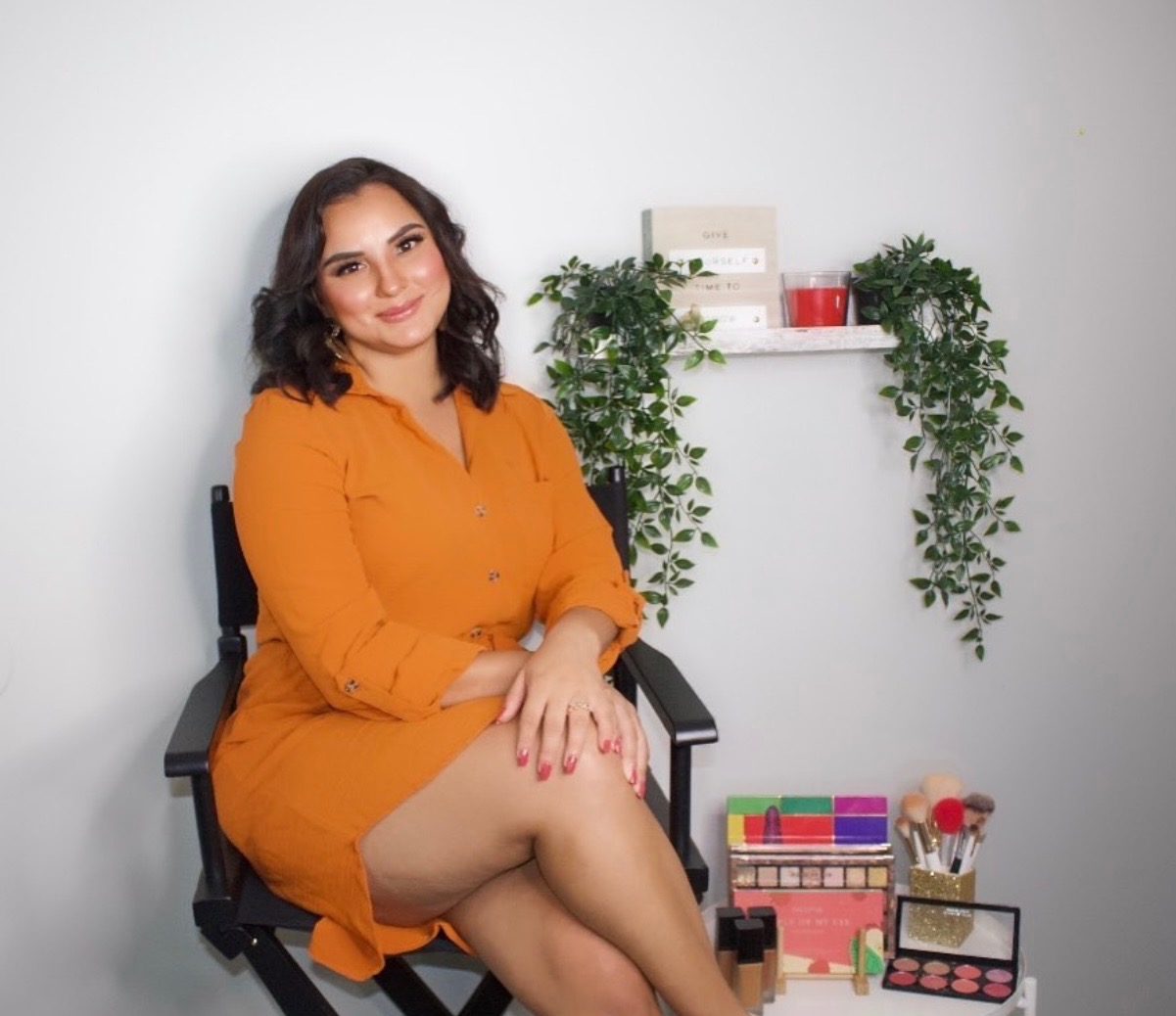 Woman with dark hair in an orange dress sitting on a chair, with shelves and plants behind her.