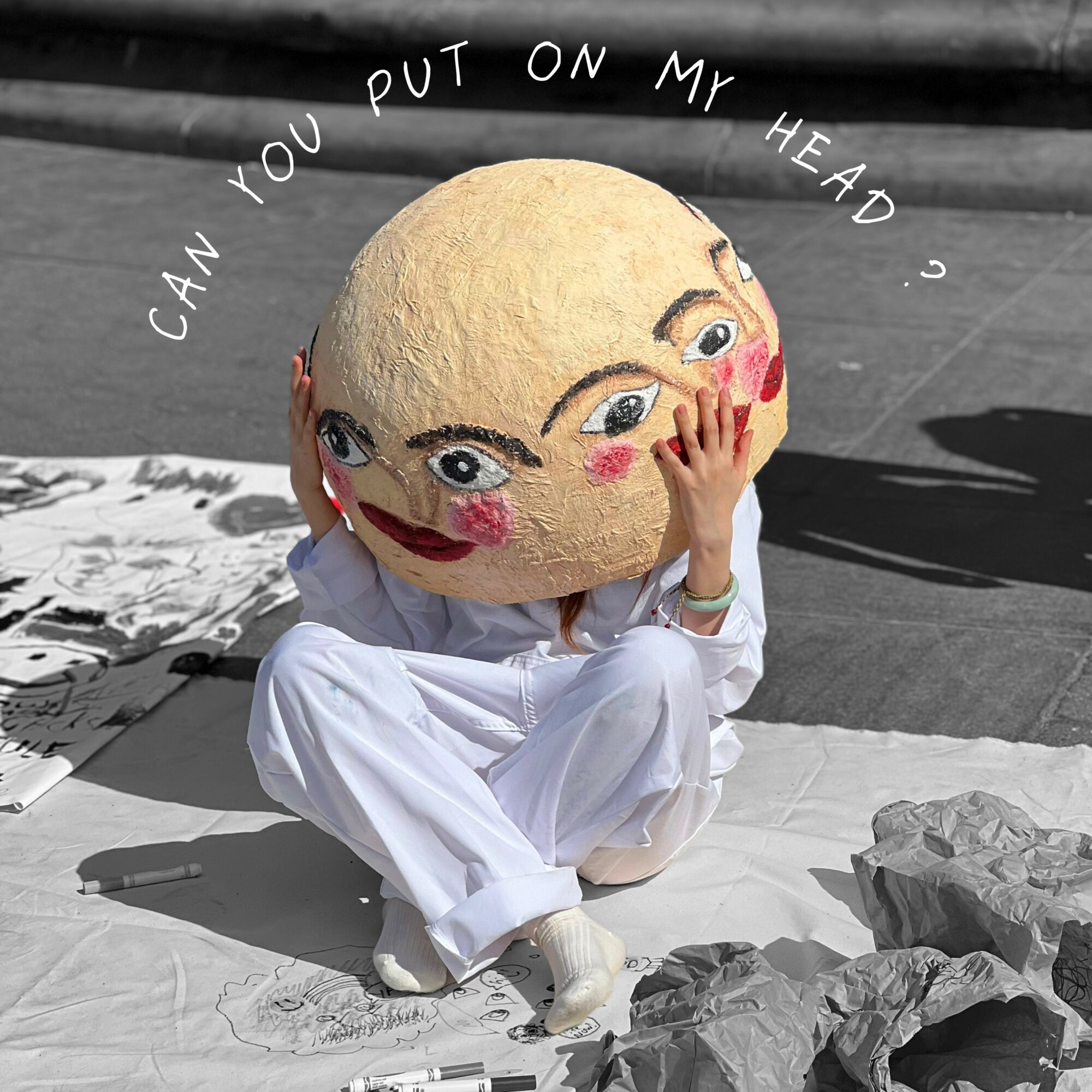 Person in white clothing with a large round head featuring painted eyes and lips, sitting on a cloth with rocks nearby.