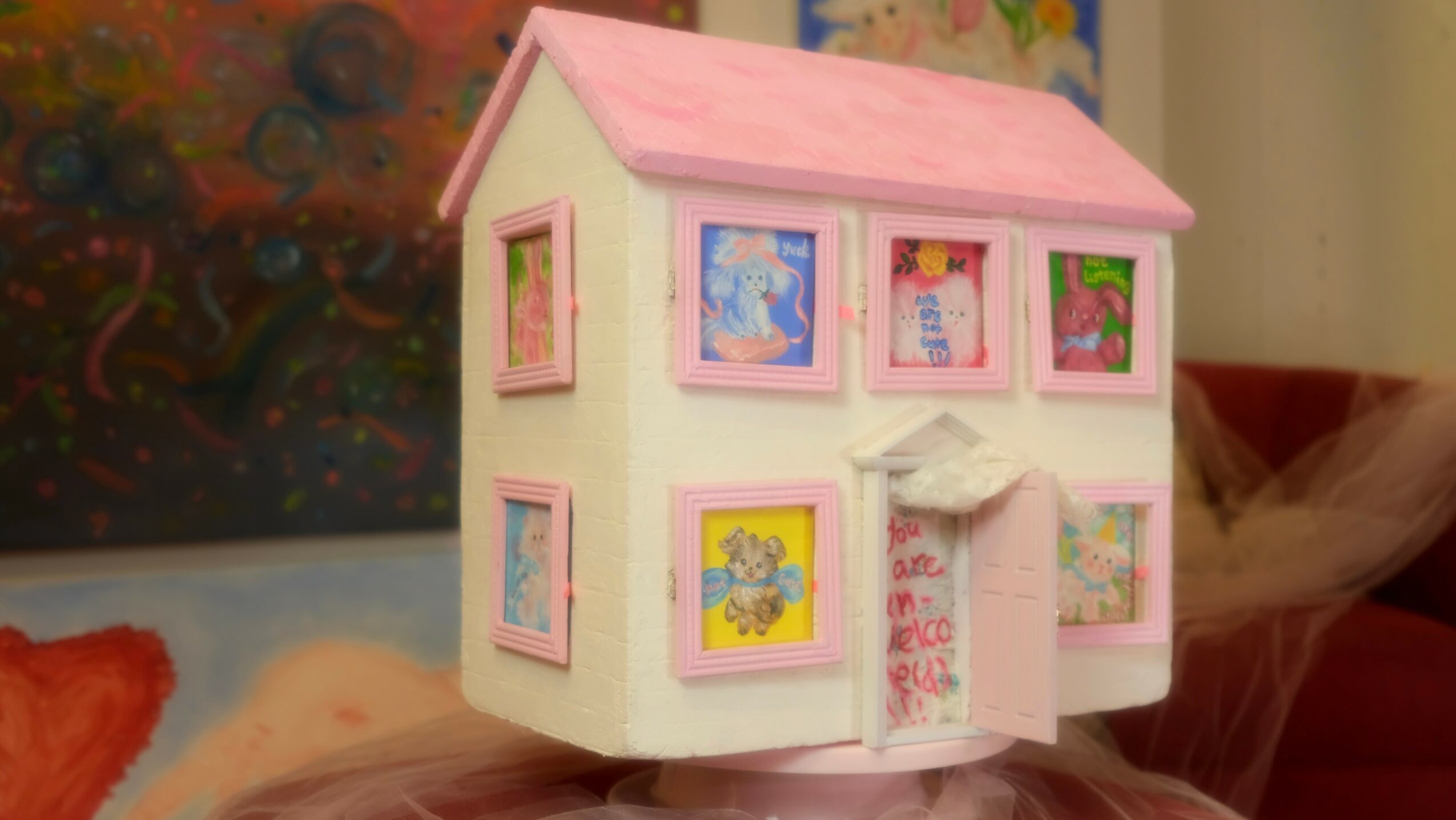 Small dollhouse with pink roof and white walls, decorated with colorful framed pictures, on a surface.