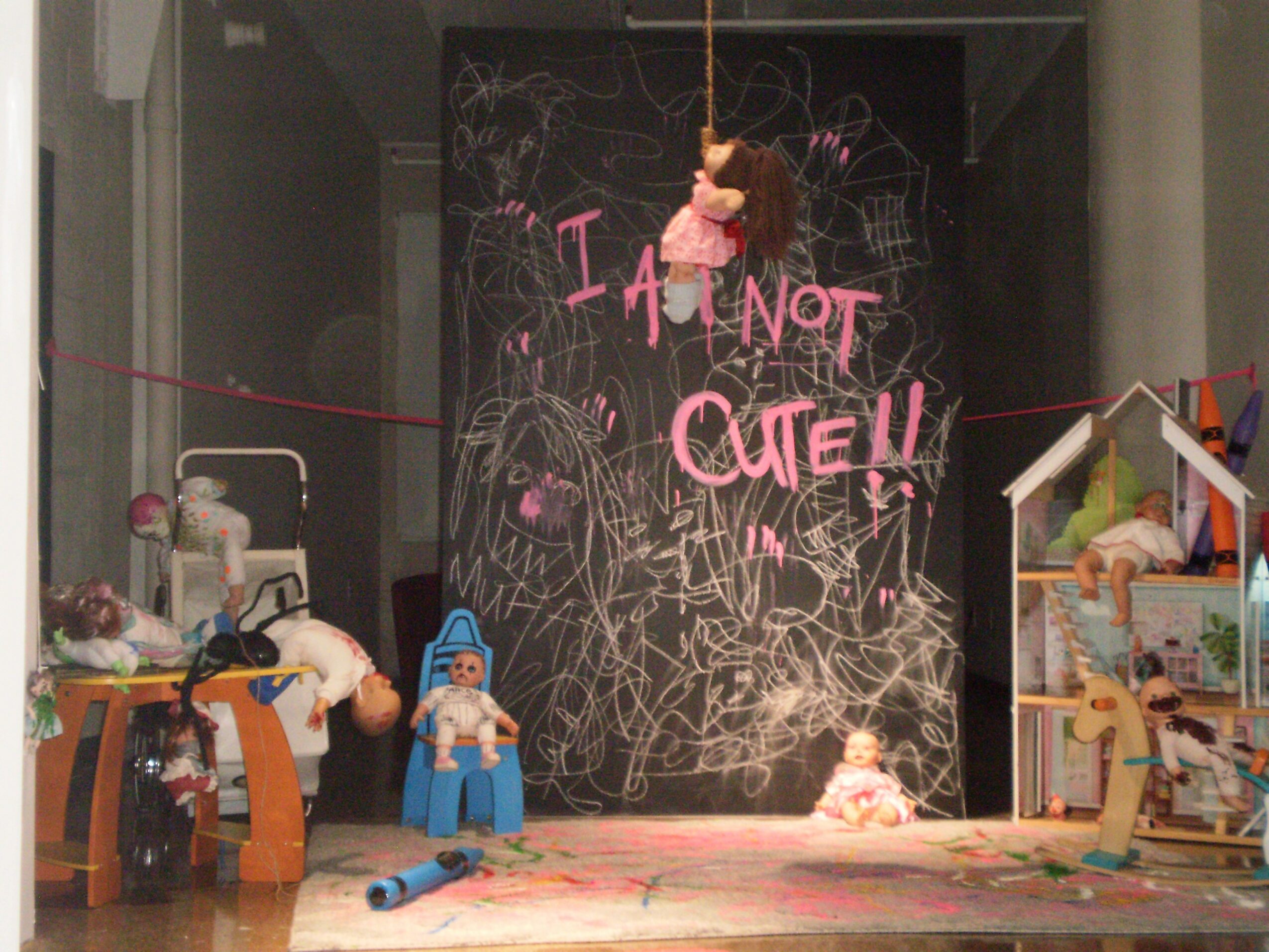 Children's toys and dolls arranged in a room with a blackboard background, some hanging and others on shelves.
