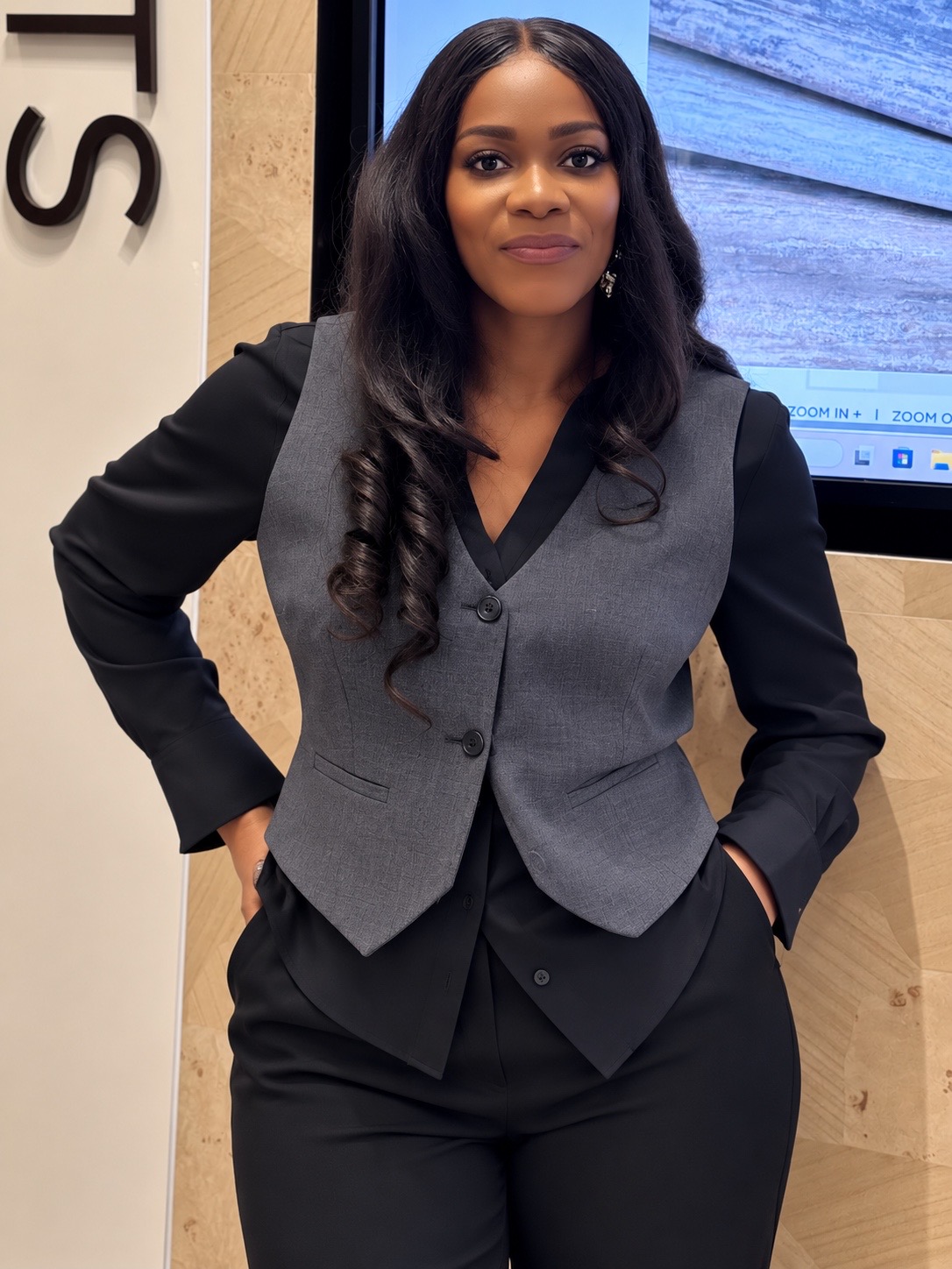 Woman with long dark hair in professional attire standing indoors with hands in pockets.