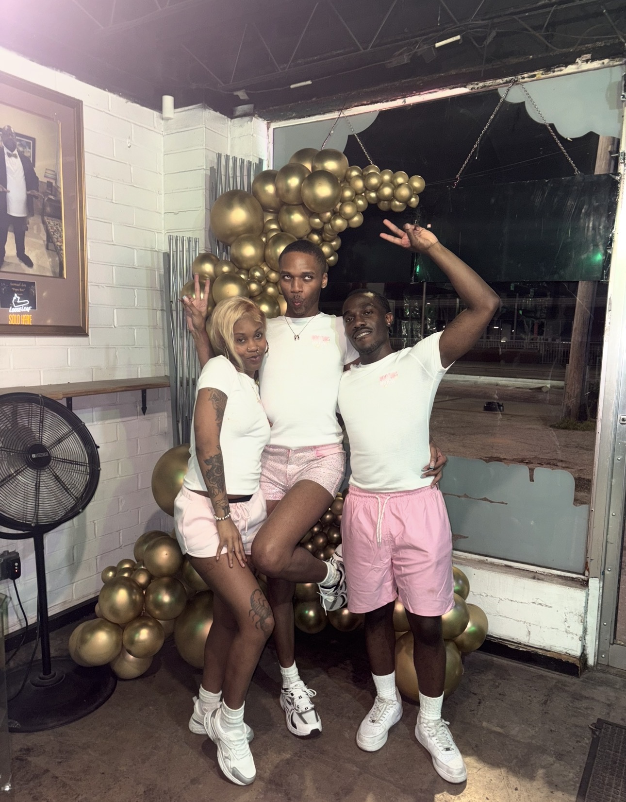 Three people standing indoors near a decorated wall with gold balloons, posing for a photo. One person is raising a hand. All are wearing casual summer outfits.