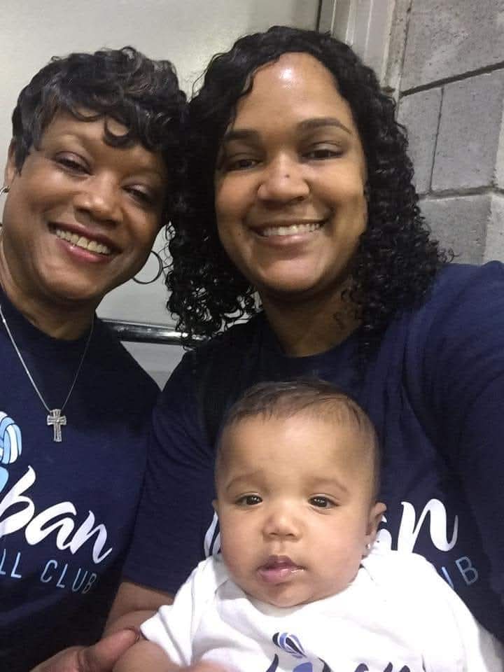 Two women and a baby smiling for a selfie indoors, with a gray wall in the background.