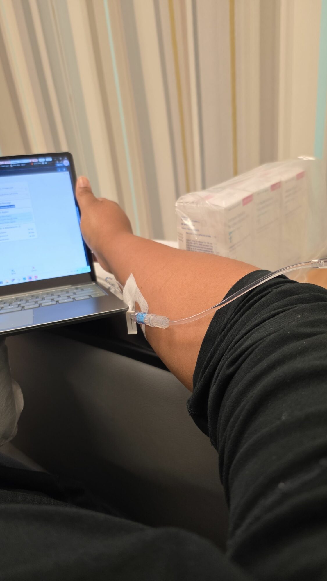 Person's arm with an IV line attached, holding a smartphone, in a medical setting.
