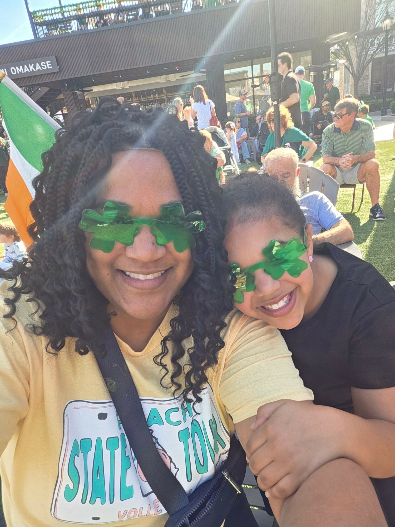 Two women smiling, wearing green shamrock glasses, in a crowded outdoor setting with people and buildings in the background.