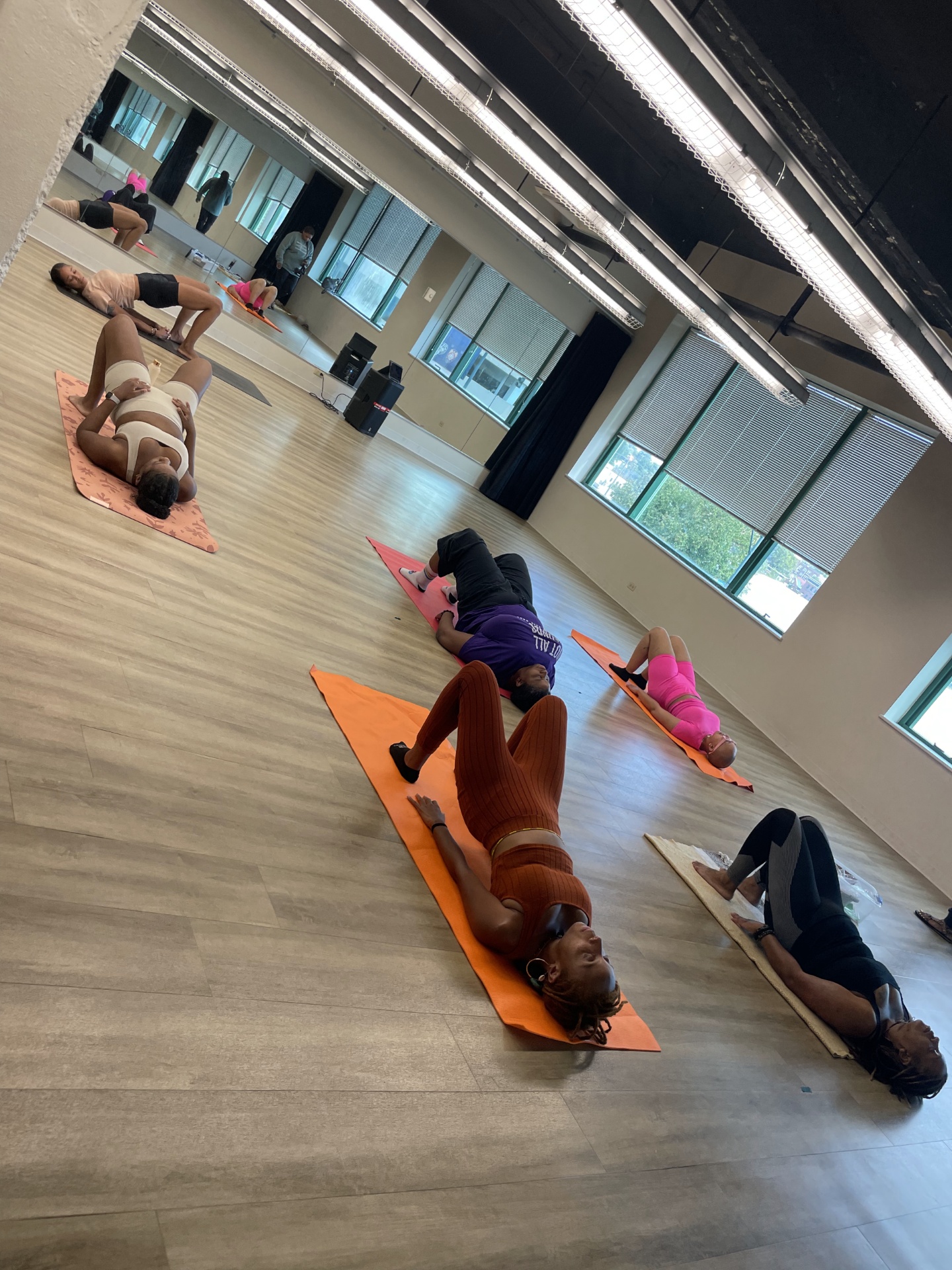 People lying on mats in a spacious room with large windows, practicing yoga or stretching exercises.