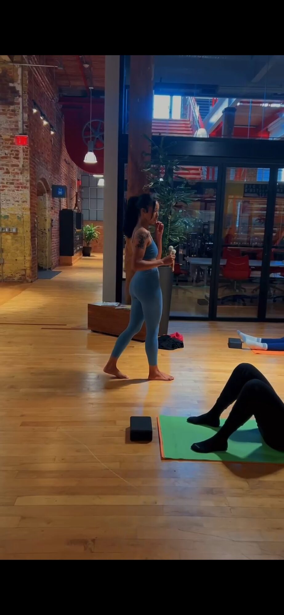 Two women in a gym, one standing and one lying on a mat, with gym equipment and large windows in the background.