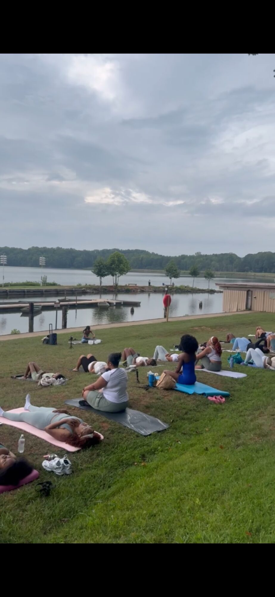 People practicing yoga and relaxing on mats by a lake on a cloudy day, with trees and a dock in the background.