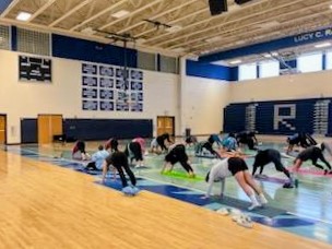 People practicing yoga on mats in a gymnasium with a wooden floor and blue walls.