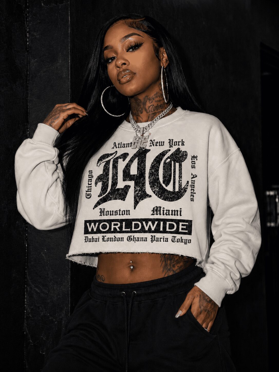 Woman with long dark hair, hoop earrings, and tattoos, wearing a white cropped sweatshirt with black text, standing against a dark background.