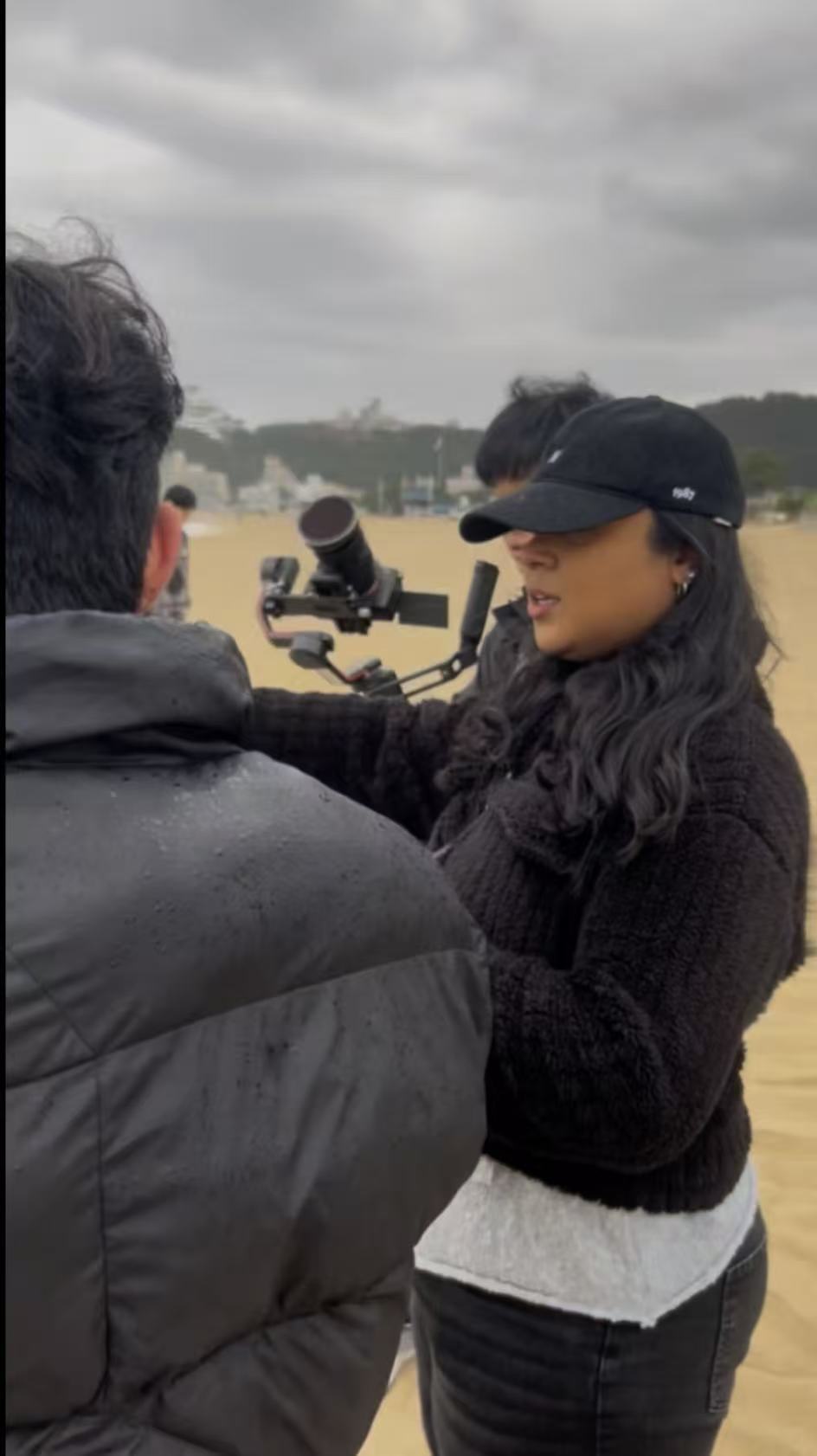 Woman with long dark hair, wearing a black cap and jacket, looks through a scope outdoors on cloudy day.