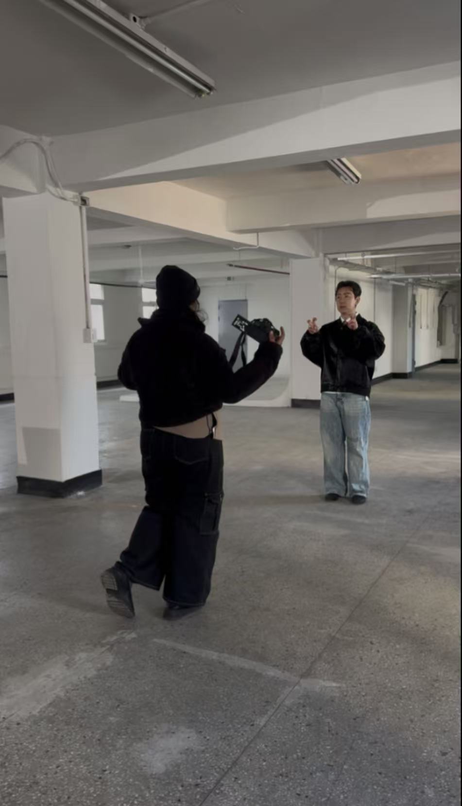 Two people standing in an indoor parking garage, one facing away and the other facing forward, with concrete pillars and ceiling lights.