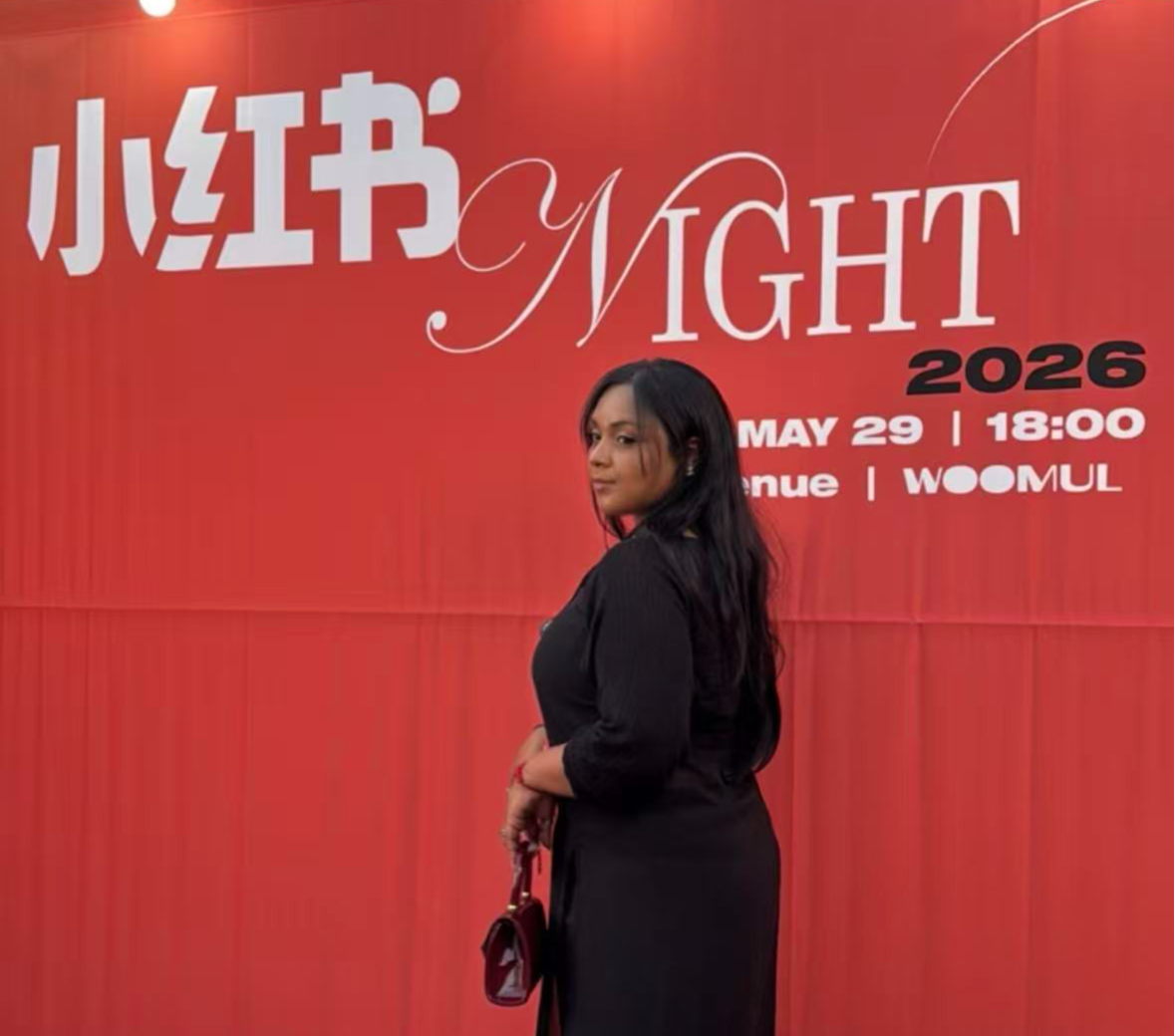 A woman with long dark hair in a black outfit stands in front of a red backdrop with event details.