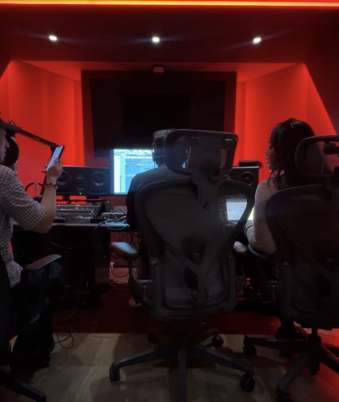 Recording studio with three people working at desks, large screen, and red lighting, viewed from behind.