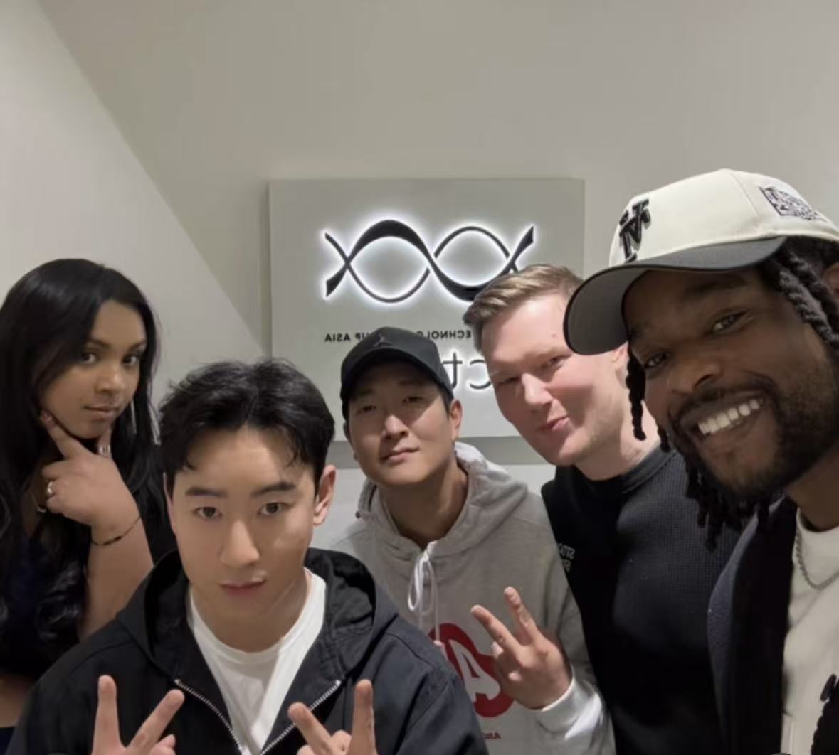 Group of five people taking a selfie indoors, smiling, with a logo on the wall behind them.
