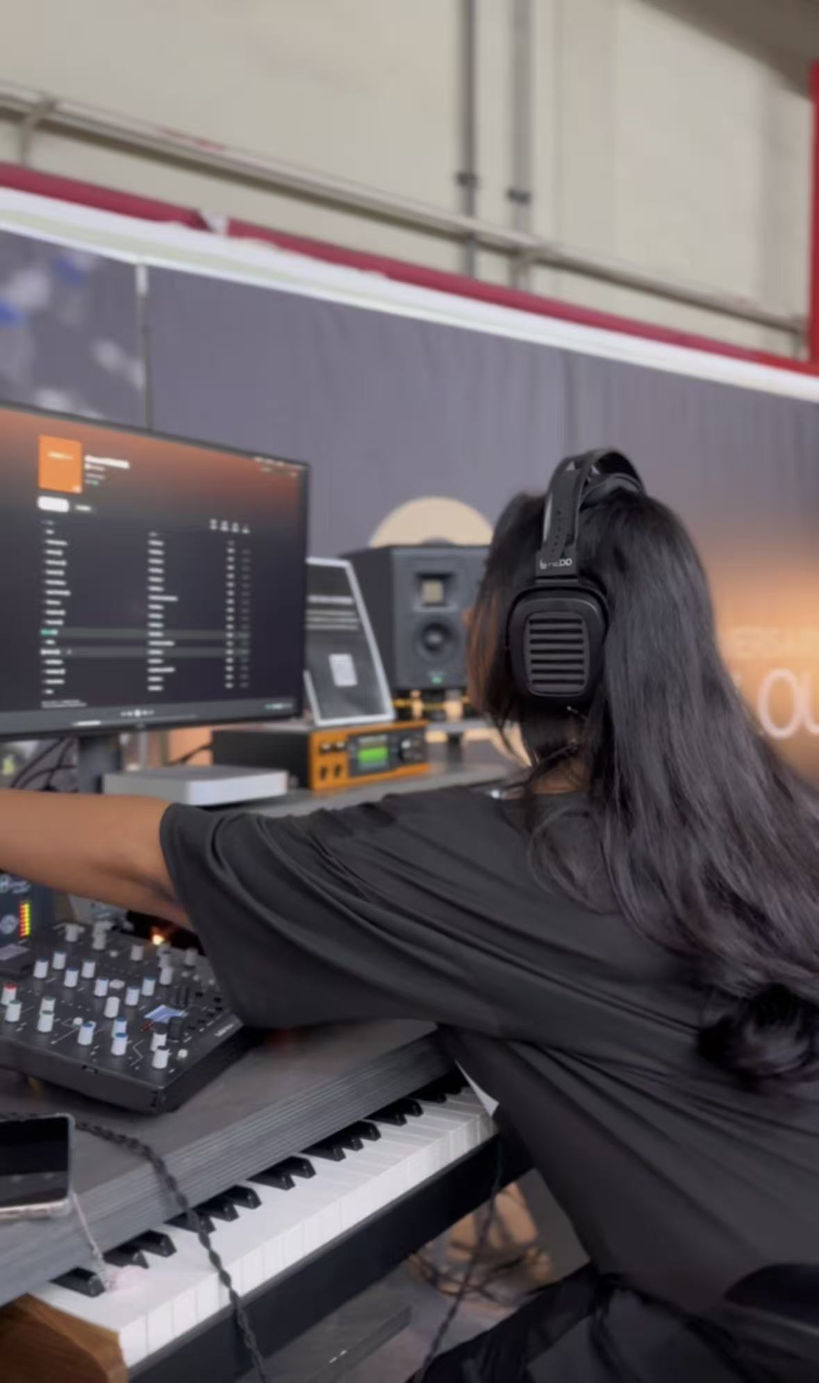 Person with long hair wearing headphones, working at a music production setup with a keyboard and computer screen.