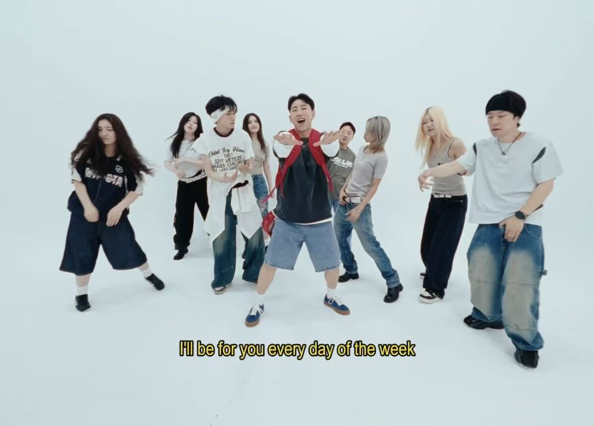 Group of eight young people dancing and singing against a plain light background, with subtitles at the bottom.