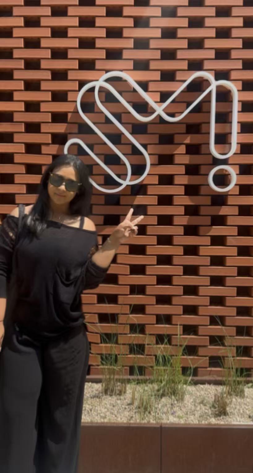 Person wearing sunglasses and a face mask making a peace sign in front of a brick wall with a white abstract sculpture.