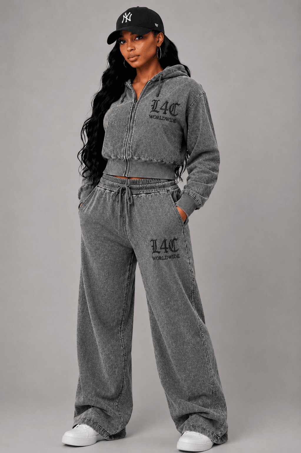 Woman in gray tracksuit with a zip-up hoodie, wide-leg pants, black cap, and white sneakers, standing with hands in pockets.