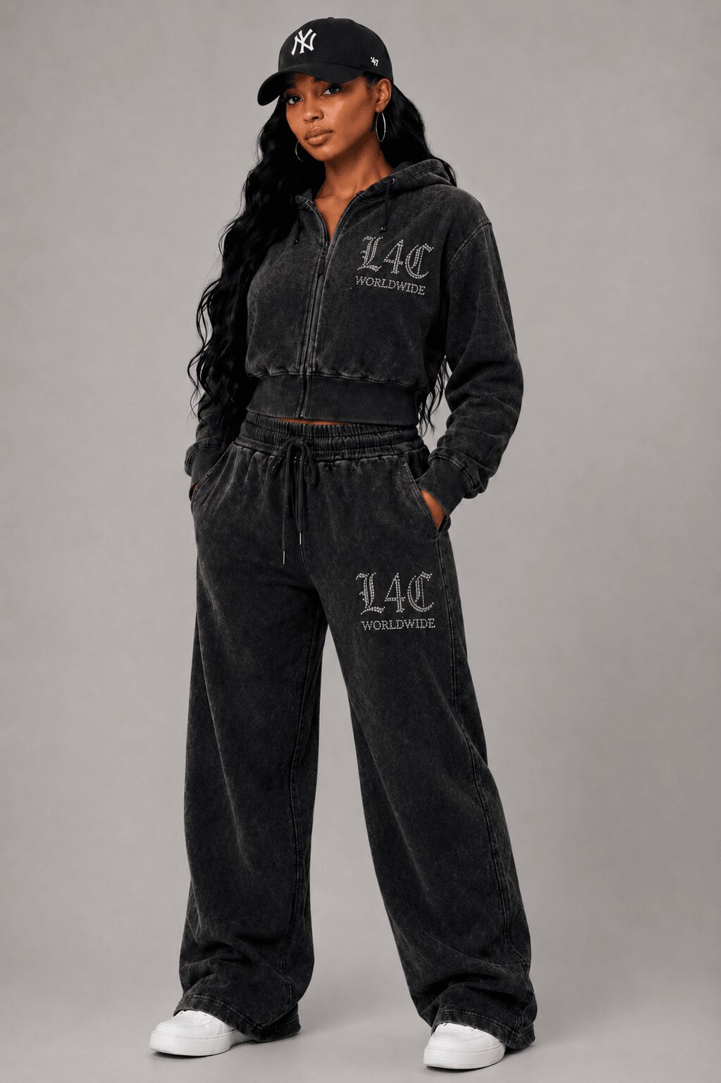 Woman in black tracksuit with white sneakers, standing against gray background, wearing a black cap, hands in pockets.