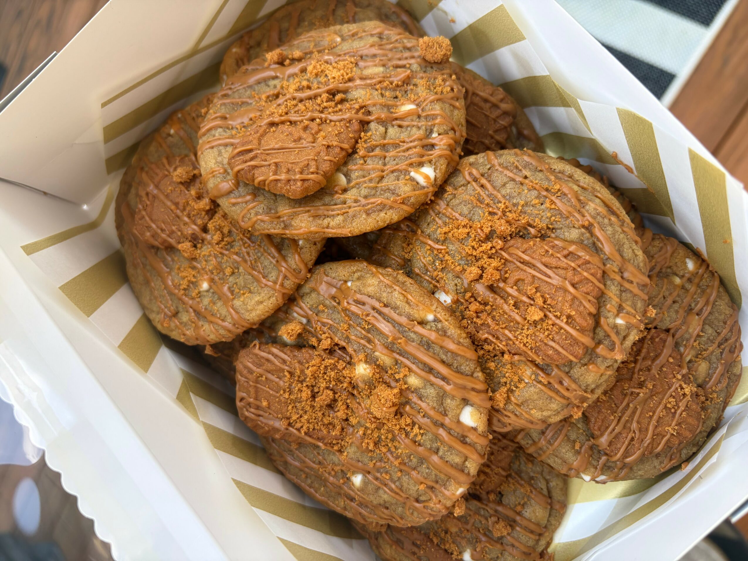 Six cookies with caramel drizzle and crushed toppings in a white and gold striped box.