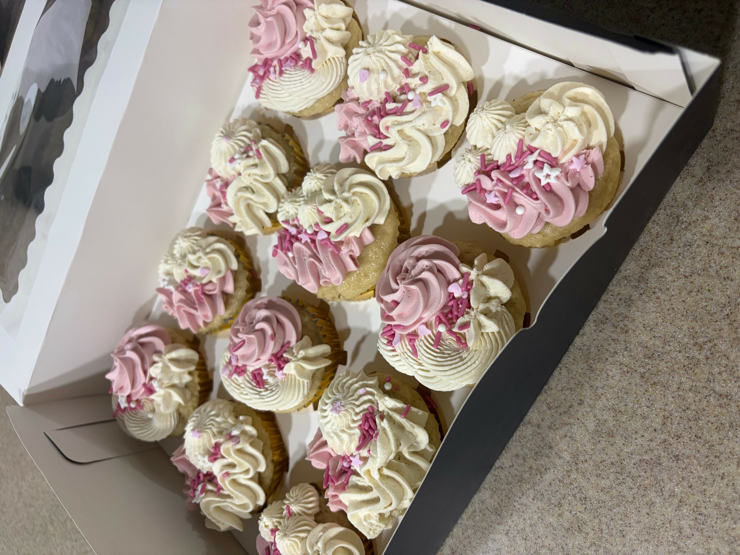 Box of cupcakes with white and pink frosting, decorated with pink sprinkles, on a gray surface.