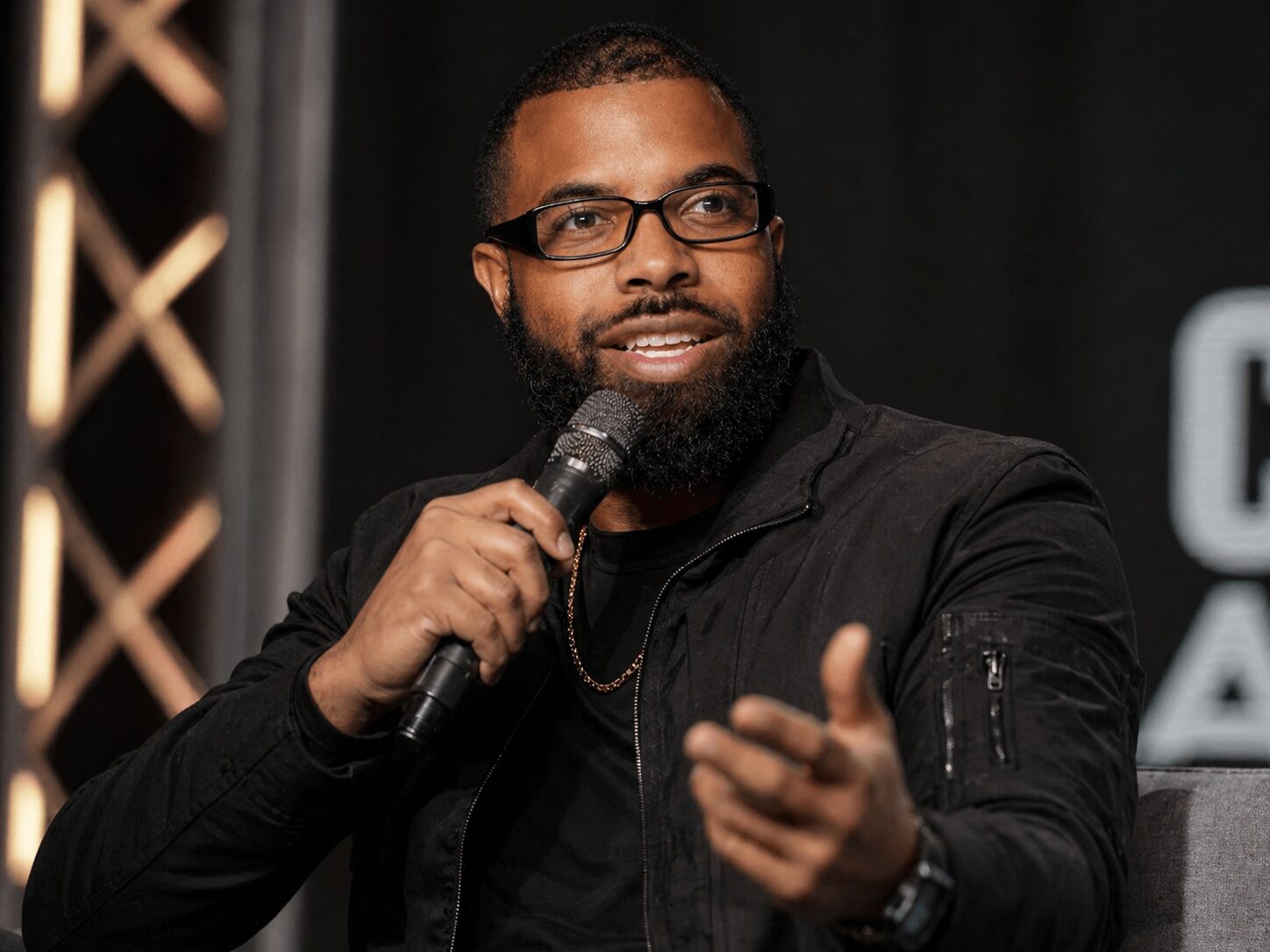 Man with glasses and beard speaking into a microphone, gesturing with his hand, wearing a black jacket, in a dark setting.