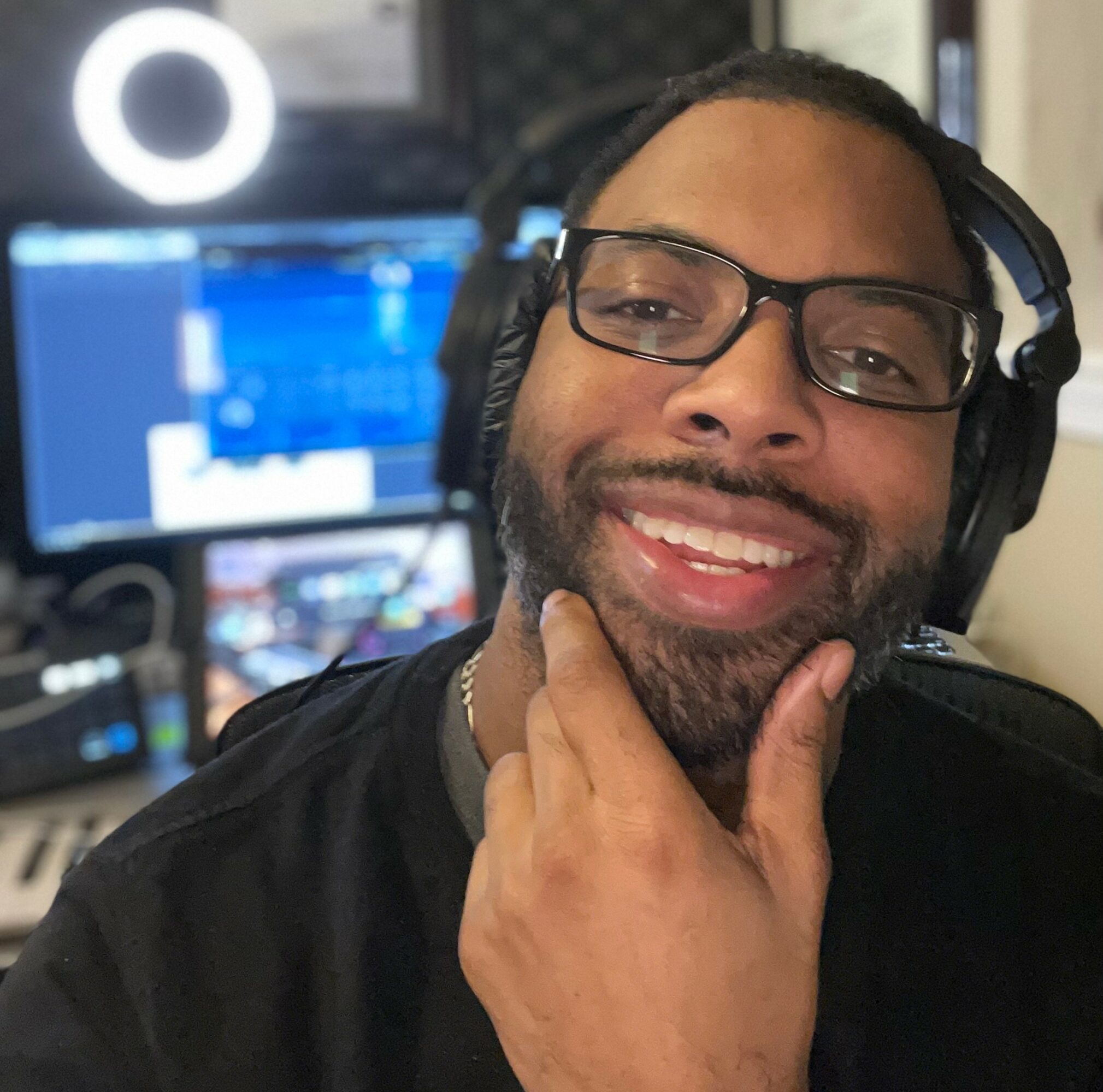Person with glasses and headphones smiling, hand on chin, in a recording studio with computer screens in background.