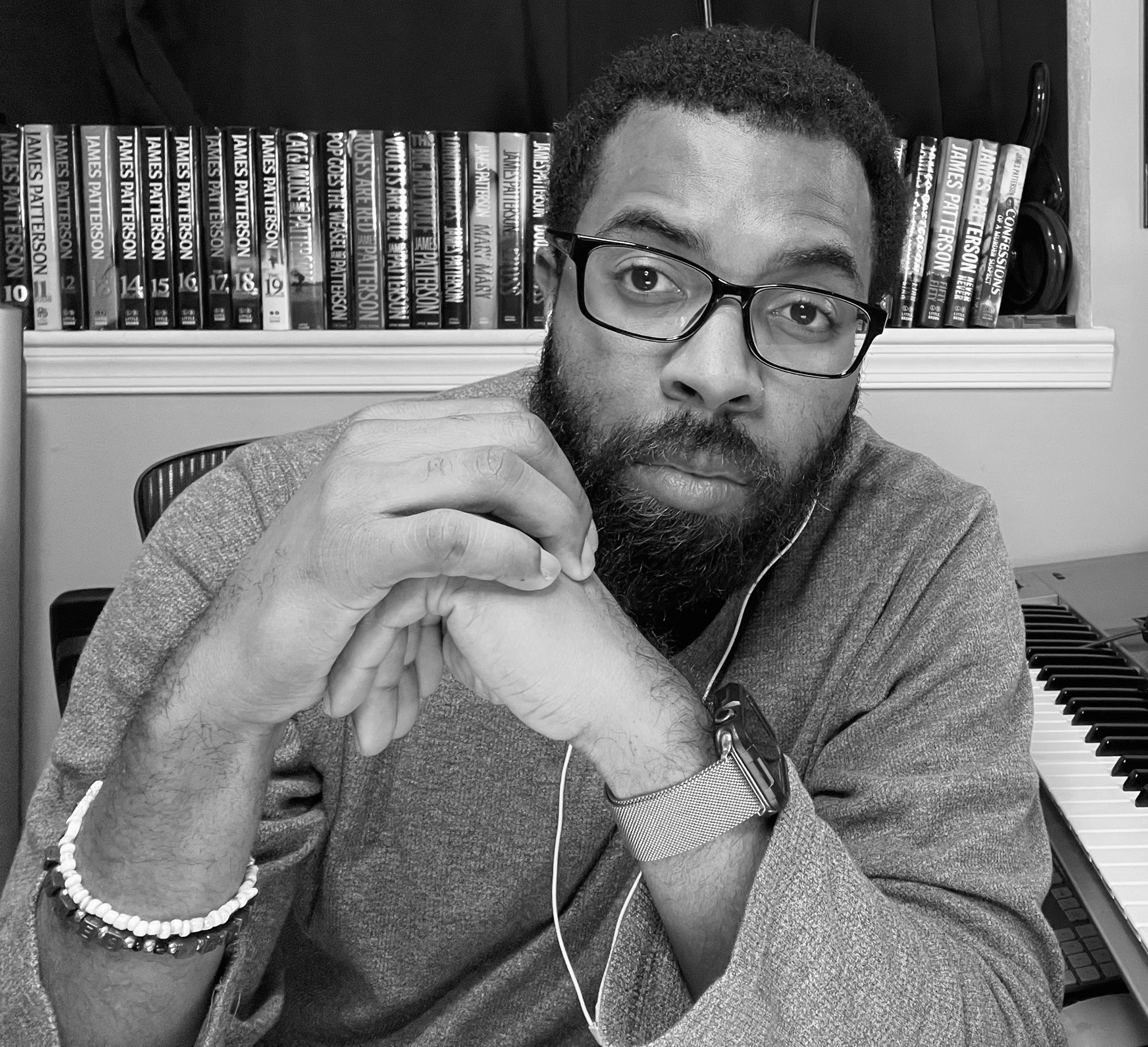 Man with glasses and beard resting his chin on hands, sitting in front of a bookshelf and piano, in black and white.