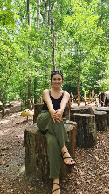 Young woman sitting on a tree stump in a forest, smiling, wearing a black top and green pants, surrounded by trees.