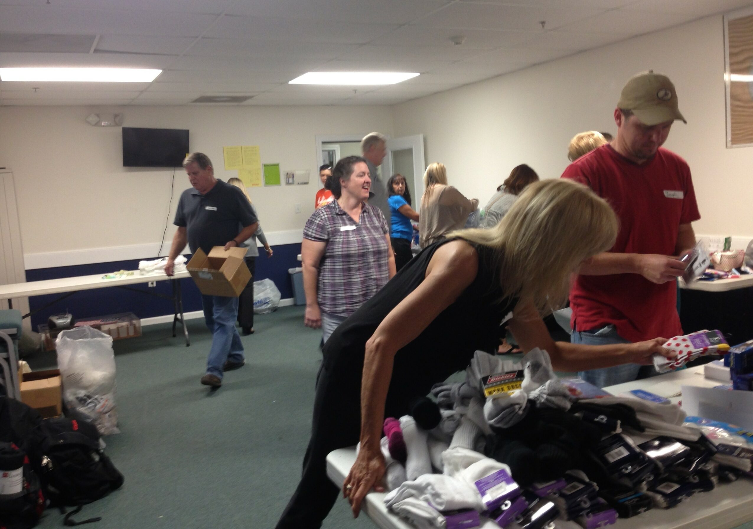 People shopping at a table with clothing and items in a room with a TV on the wall.