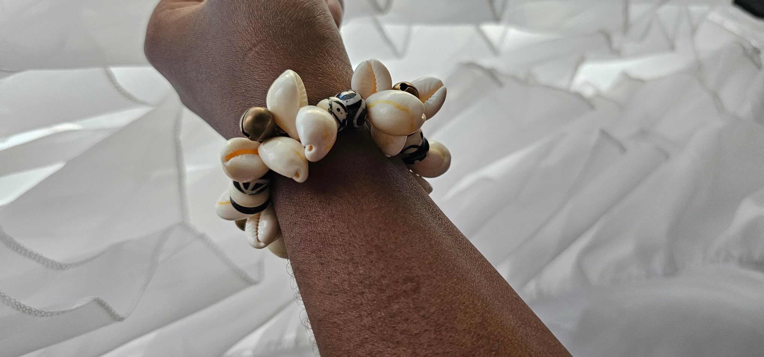 Person's wrist wearing a shell bracelet with white and black shells, against a white fabric background.