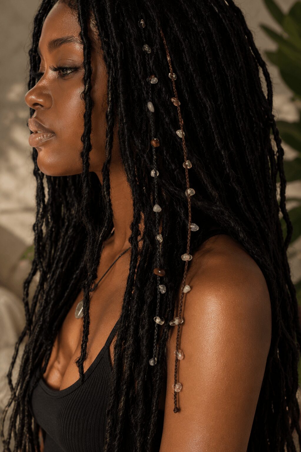 Side profile of a woman with long, decorated dreadlocks and a black top, outdoors with greenery in background.