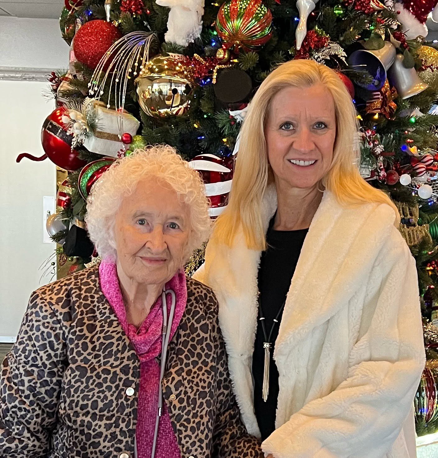 Two women standing in front of a decorated Christmas tree, smiling, one elderly with curly white hair, the other middle-aged with long blonde hair.