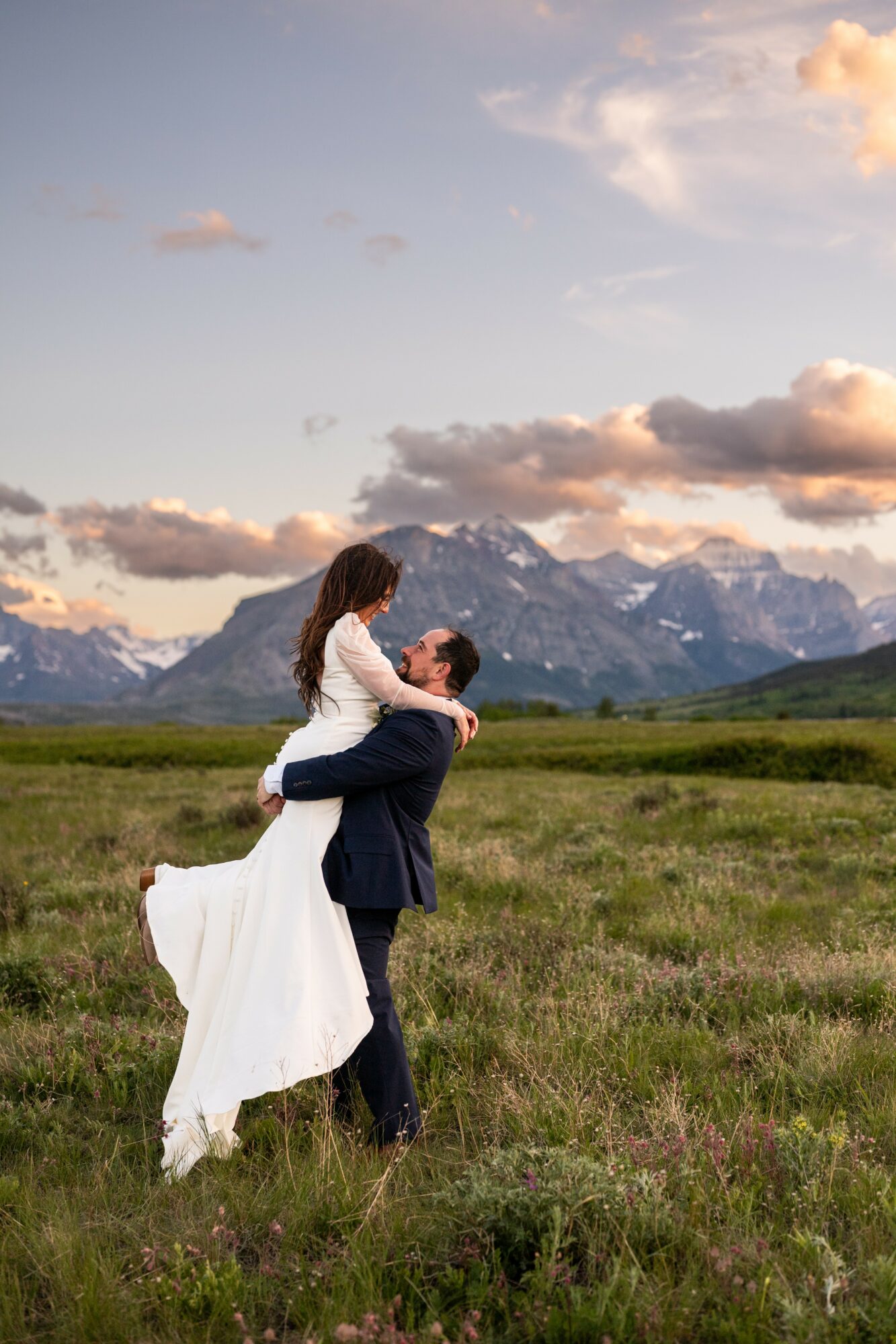 A couple in formal attire standing in a grassy field with mountains and a partly cloudy sky in the background.