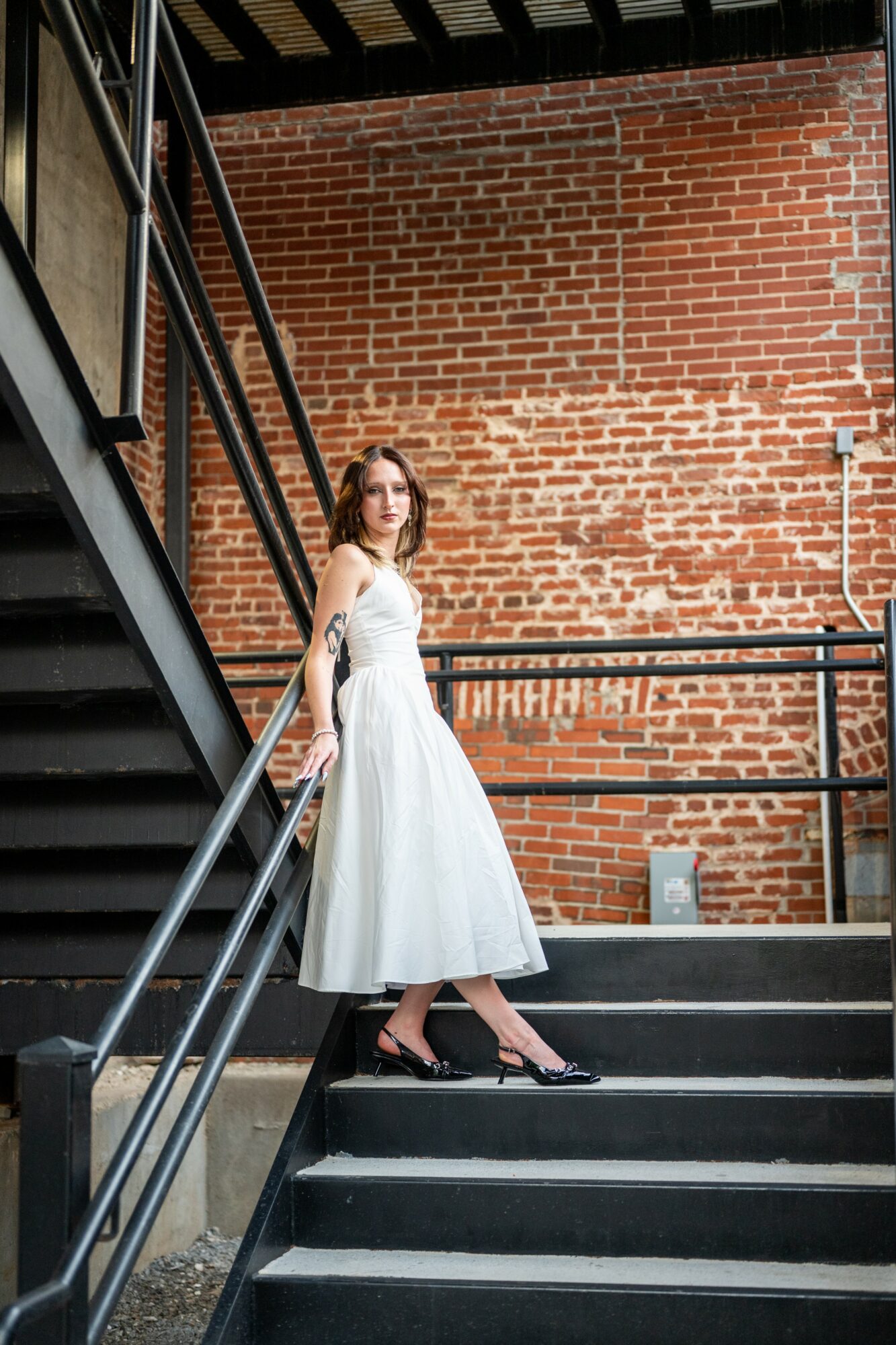 Woman in white dress standing on stairs against brick wall, looking at camera, with metal railing, daytime.