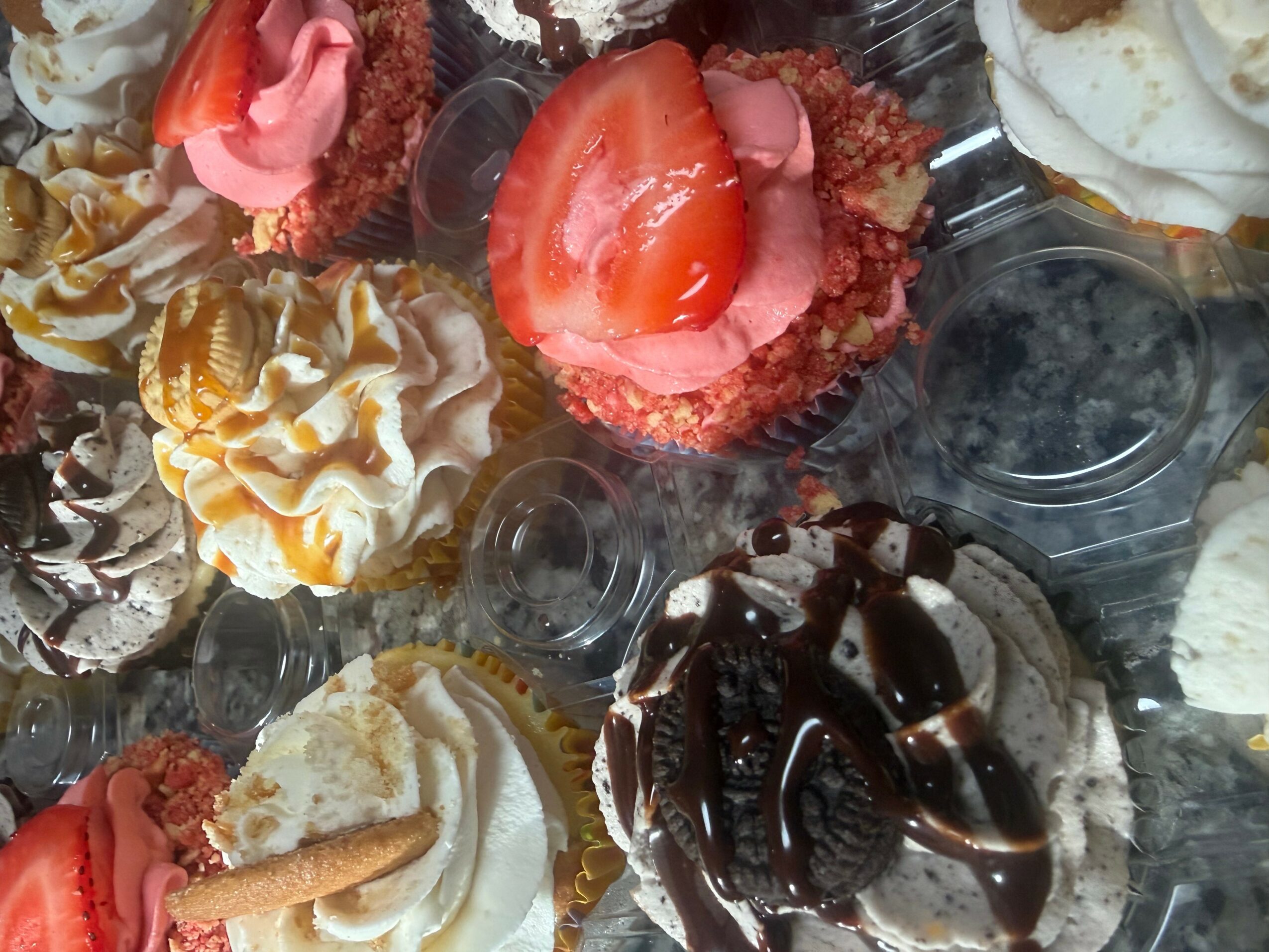 Assorted cupcakes with various toppings, including strawberries, chocolate drizzle, and whipped cream, arranged on a tray.