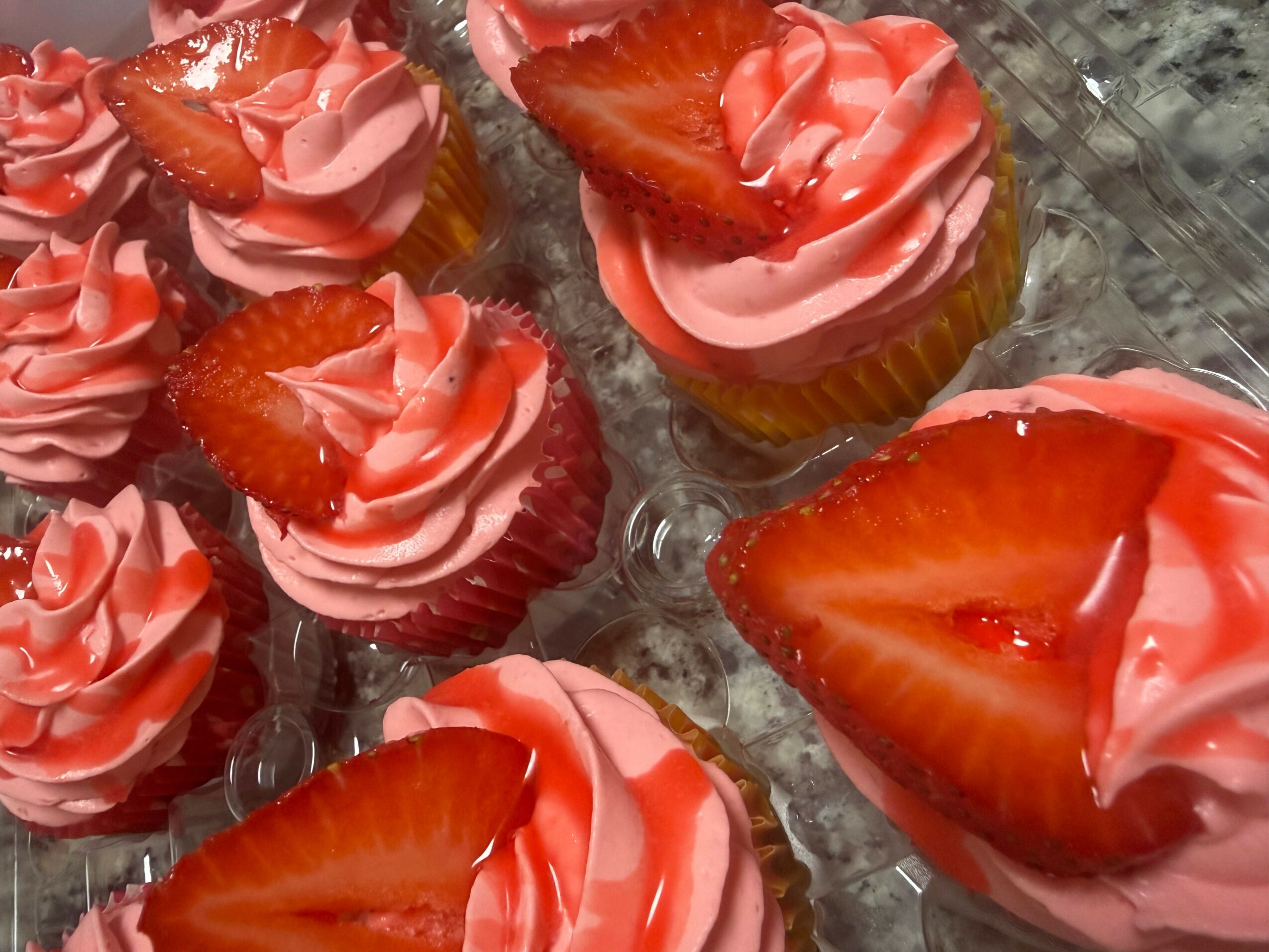 Six cupcakes with pink frosting and strawberry slices on top, arranged on a clear plastic tray.