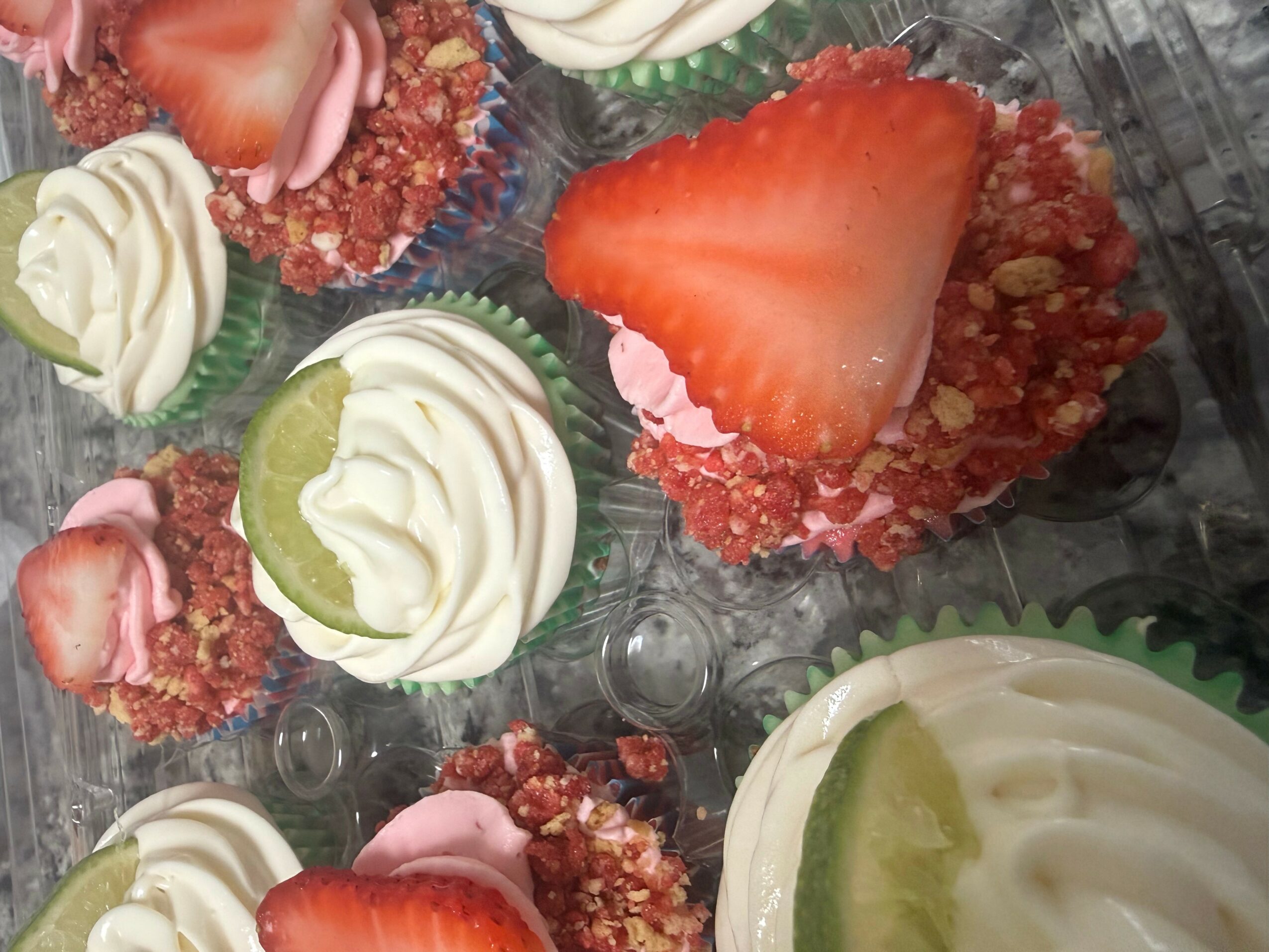 Cupcakes topped with strawberries, whipped cream, and green fruit slices, arranged in a plastic container.