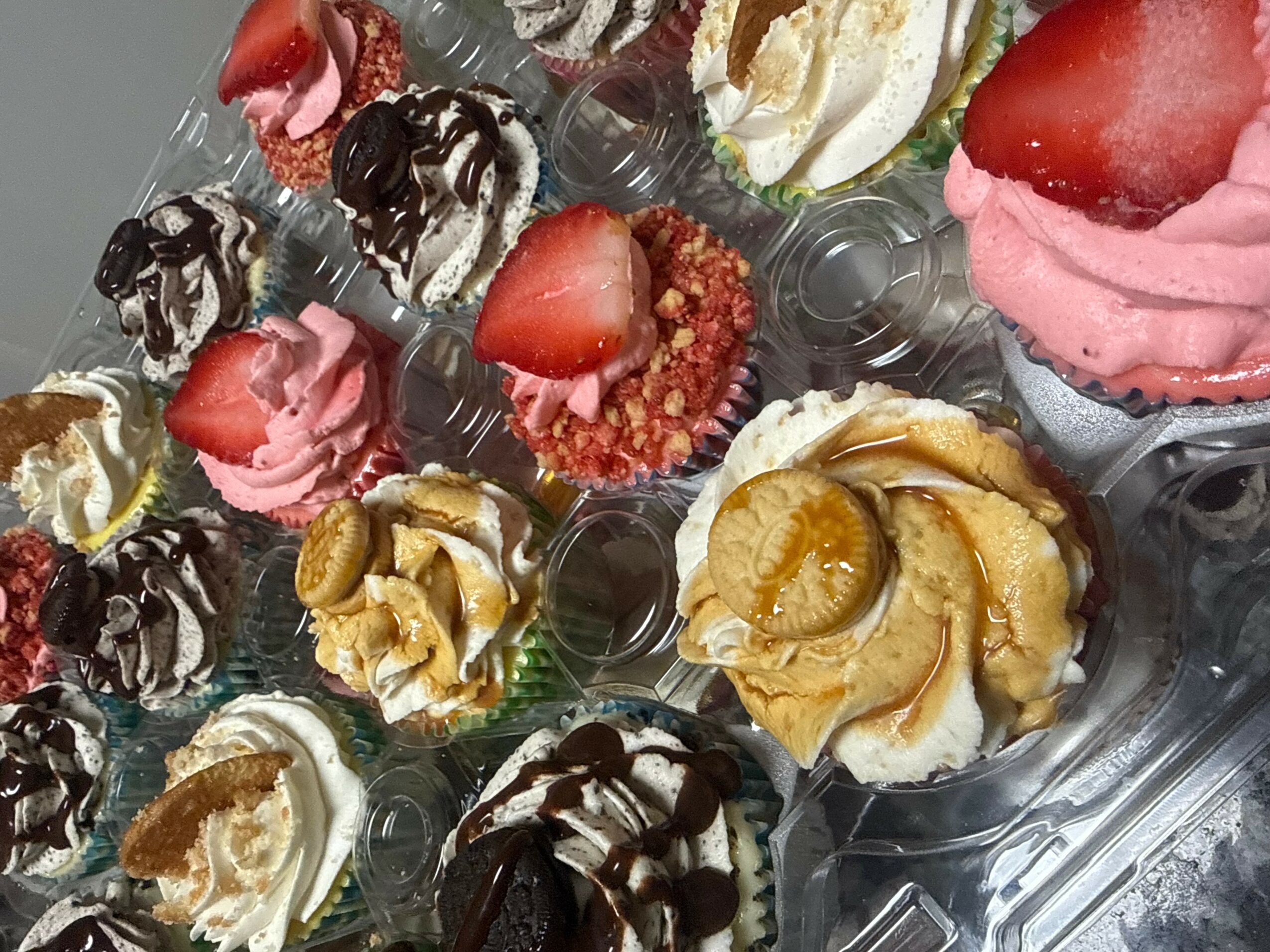 Assorted cupcakes with various toppings, including strawberries, chocolate, and cream, arranged in a plastic container.