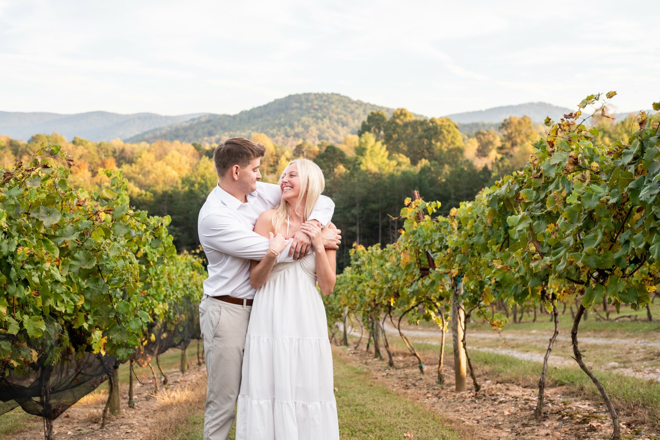 A couple stands in a vineyard with green and yellow foliage, mountains in the background, under a cloudy sky.