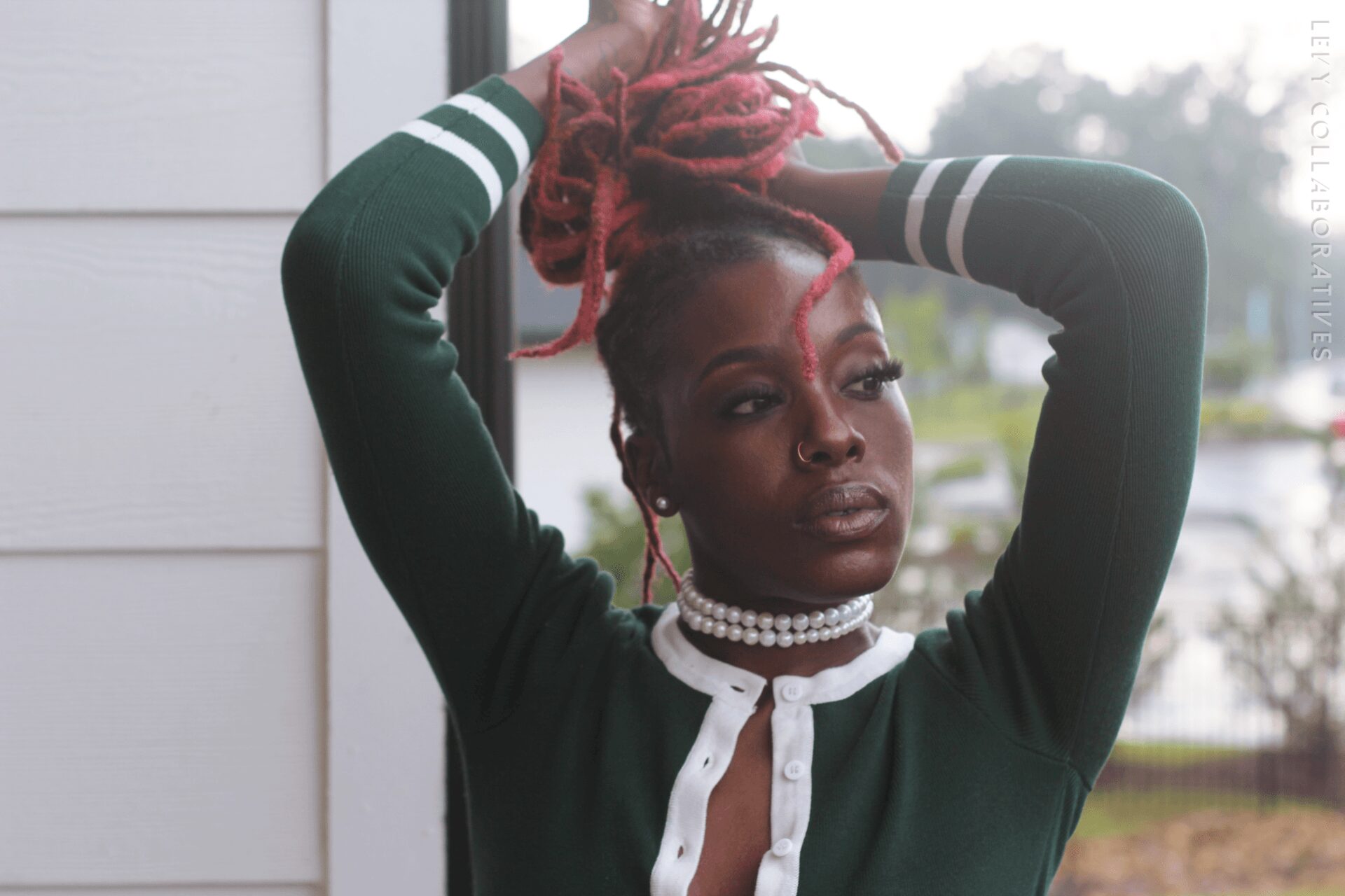 Woman with pink dreadlocks and dark lipstick poses outdoors, wearing a green and white sports jacket and pearl necklace.