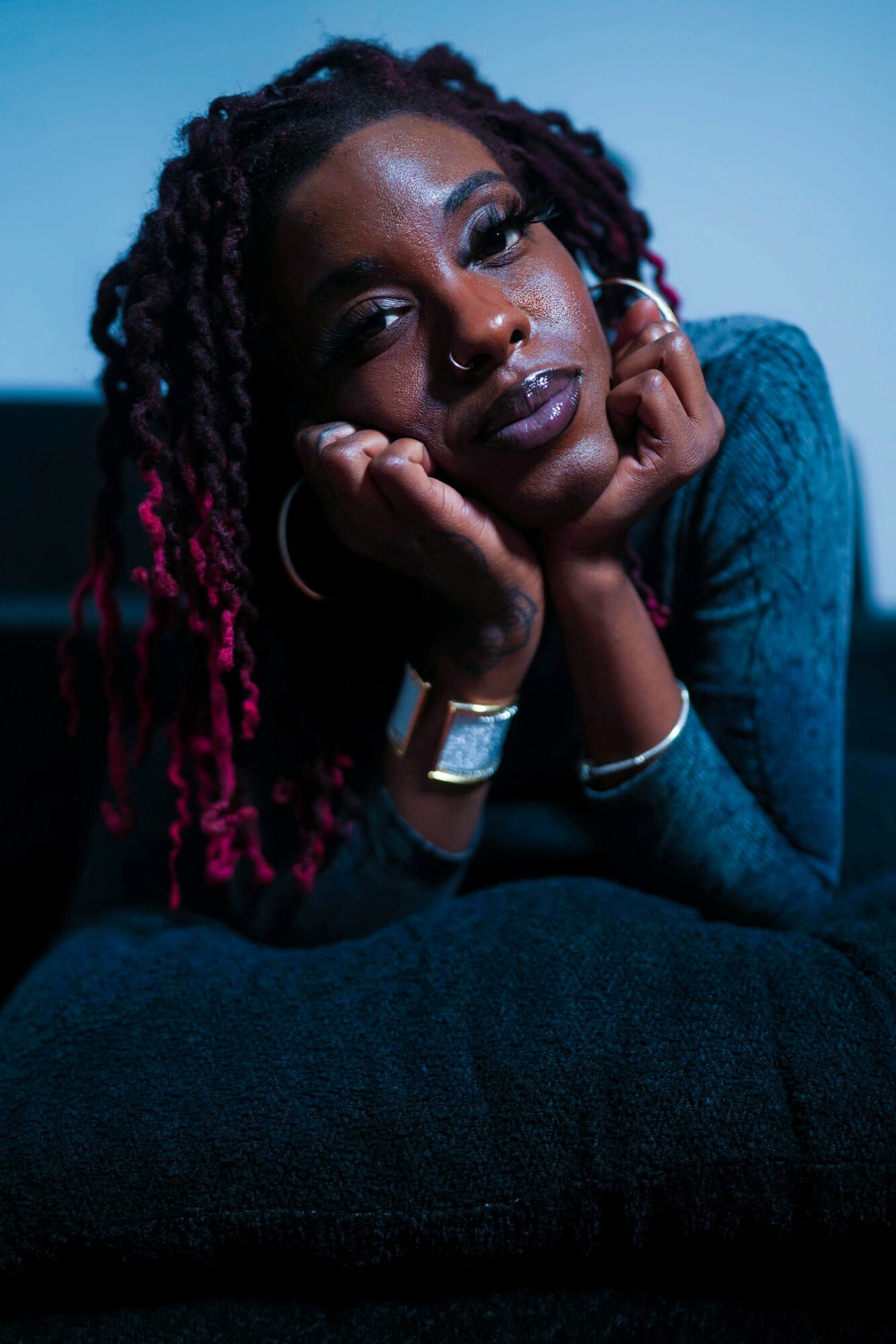 Woman with dreadlocks resting her face on her hands, looking at the camera, dark background, blue lighting.