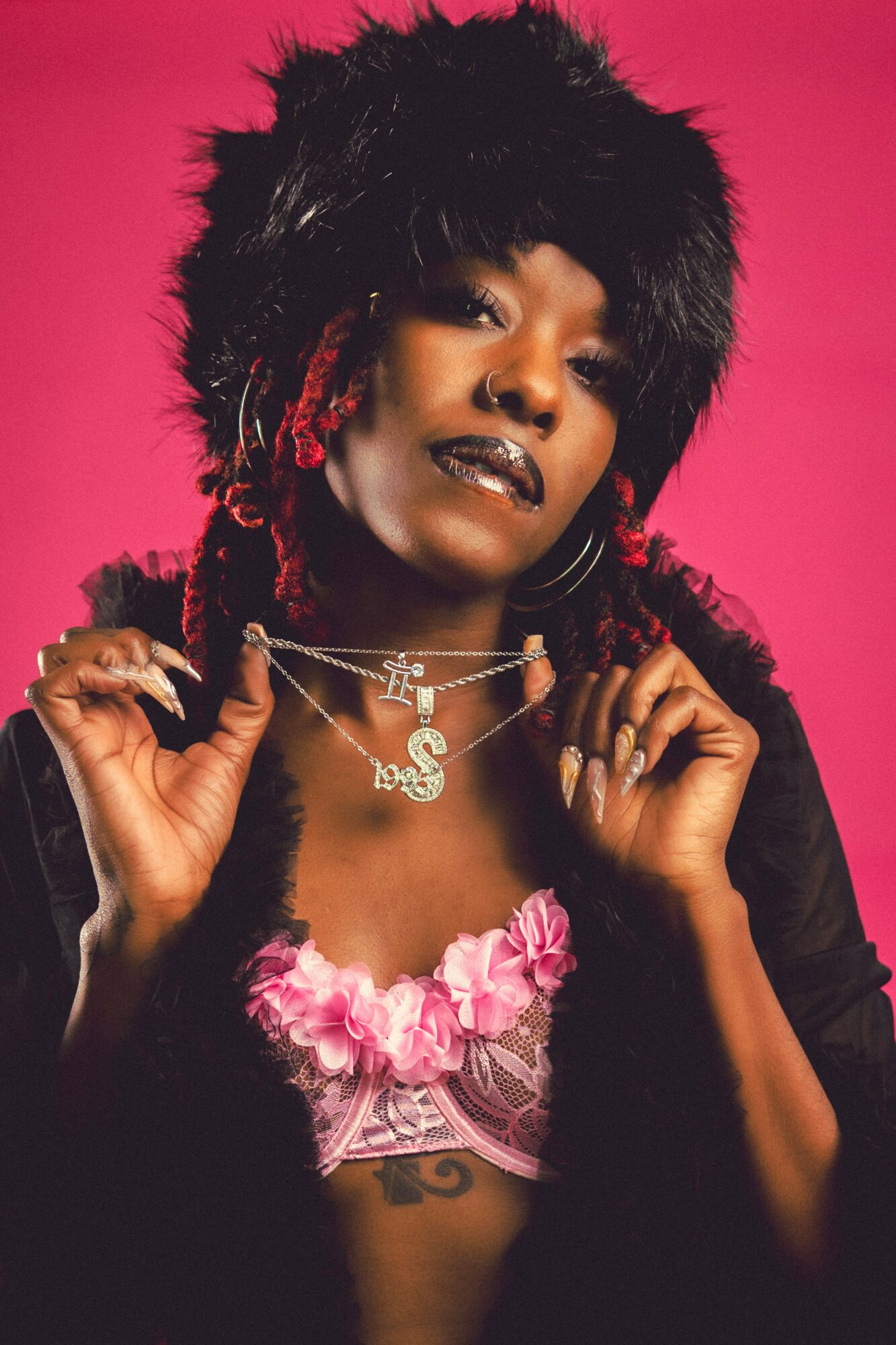 Woman with short black hair, dark lipstick, jewelry, and pink clothing against a pink background.
