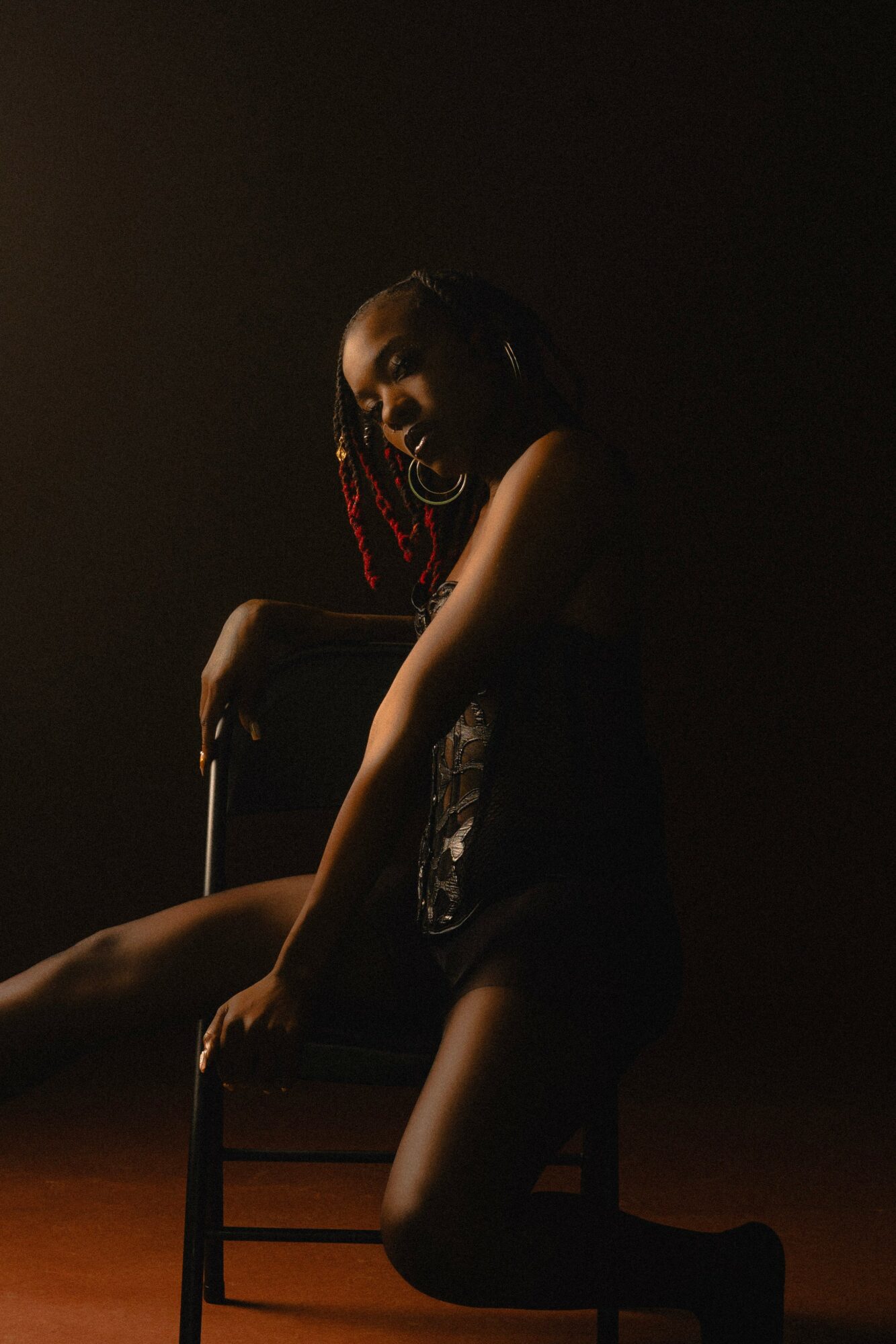 Person sitting on a chair in a dark room, with light highlighting their face and body, and red hair accessories.