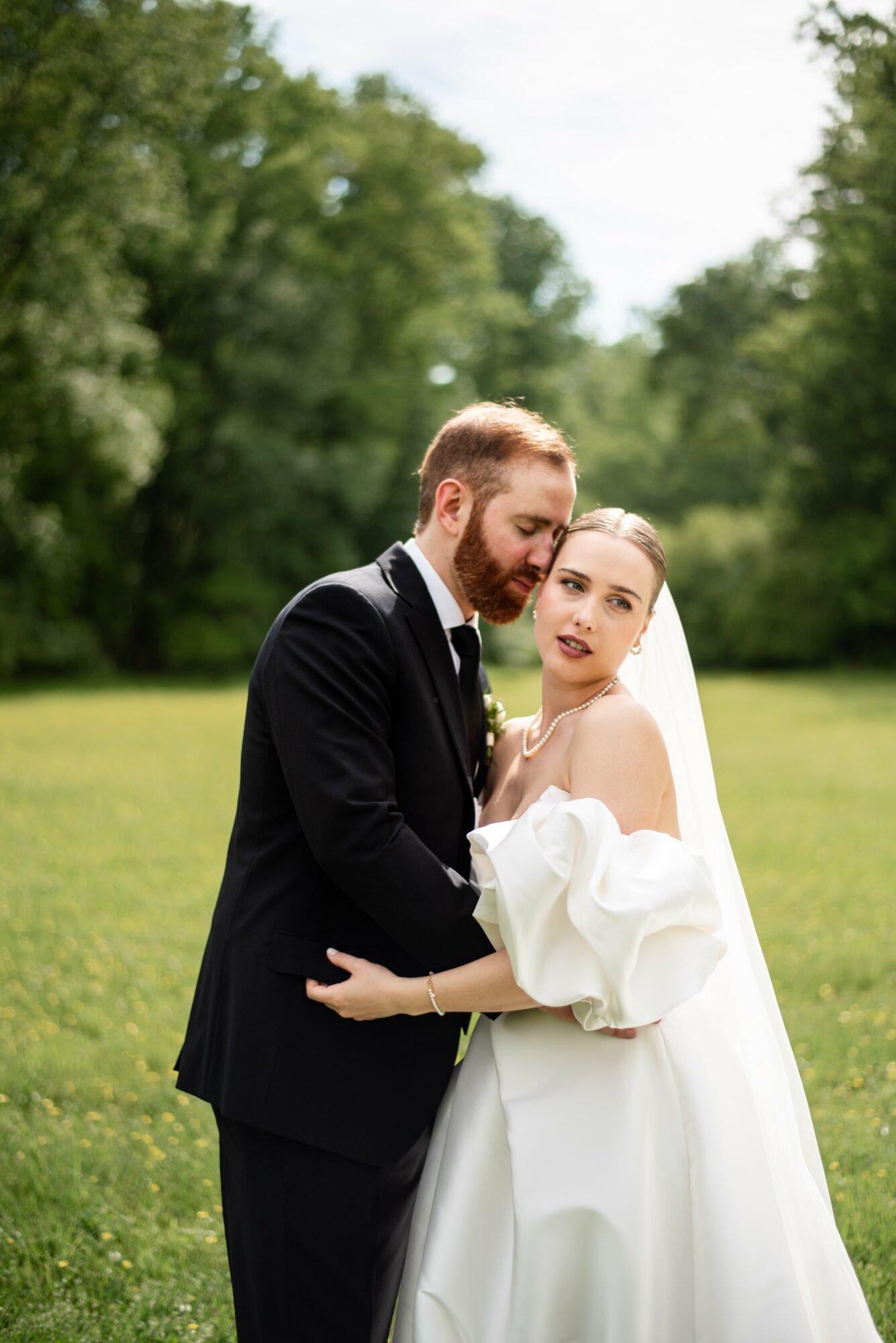 A bride and groom embrace outdoors in a green park, with trees and sky in the background.