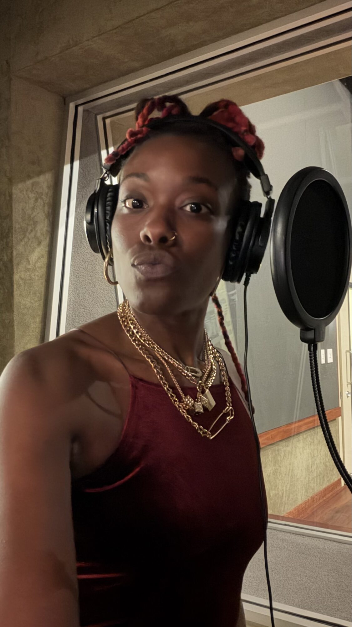 Woman in recording studio wearing headphones, red top, and layered gold necklaces, looking at camera with a serious expression.