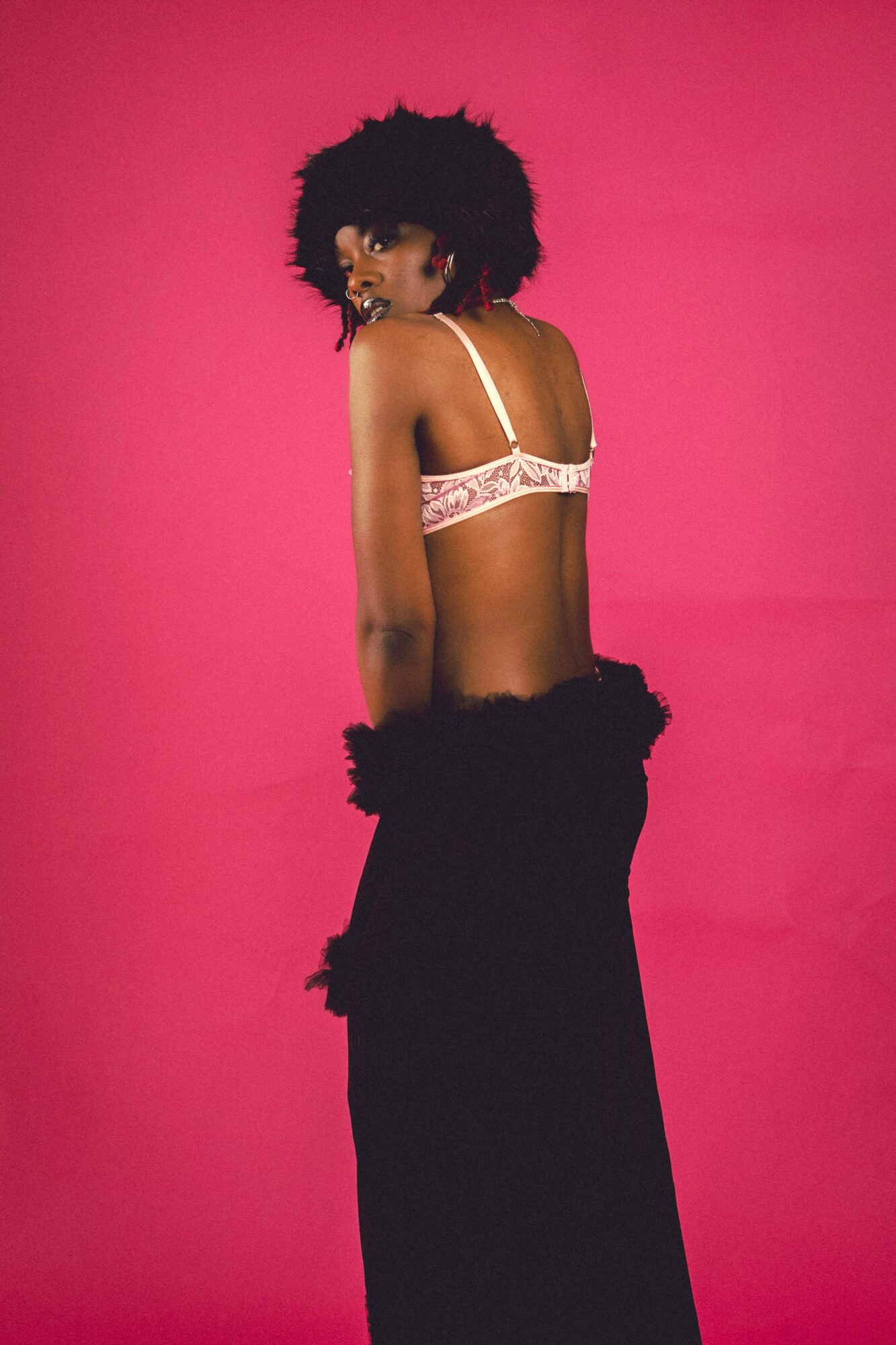 Woman with curly hair in lingerie and a black skirt against a pink background, turned sideways.