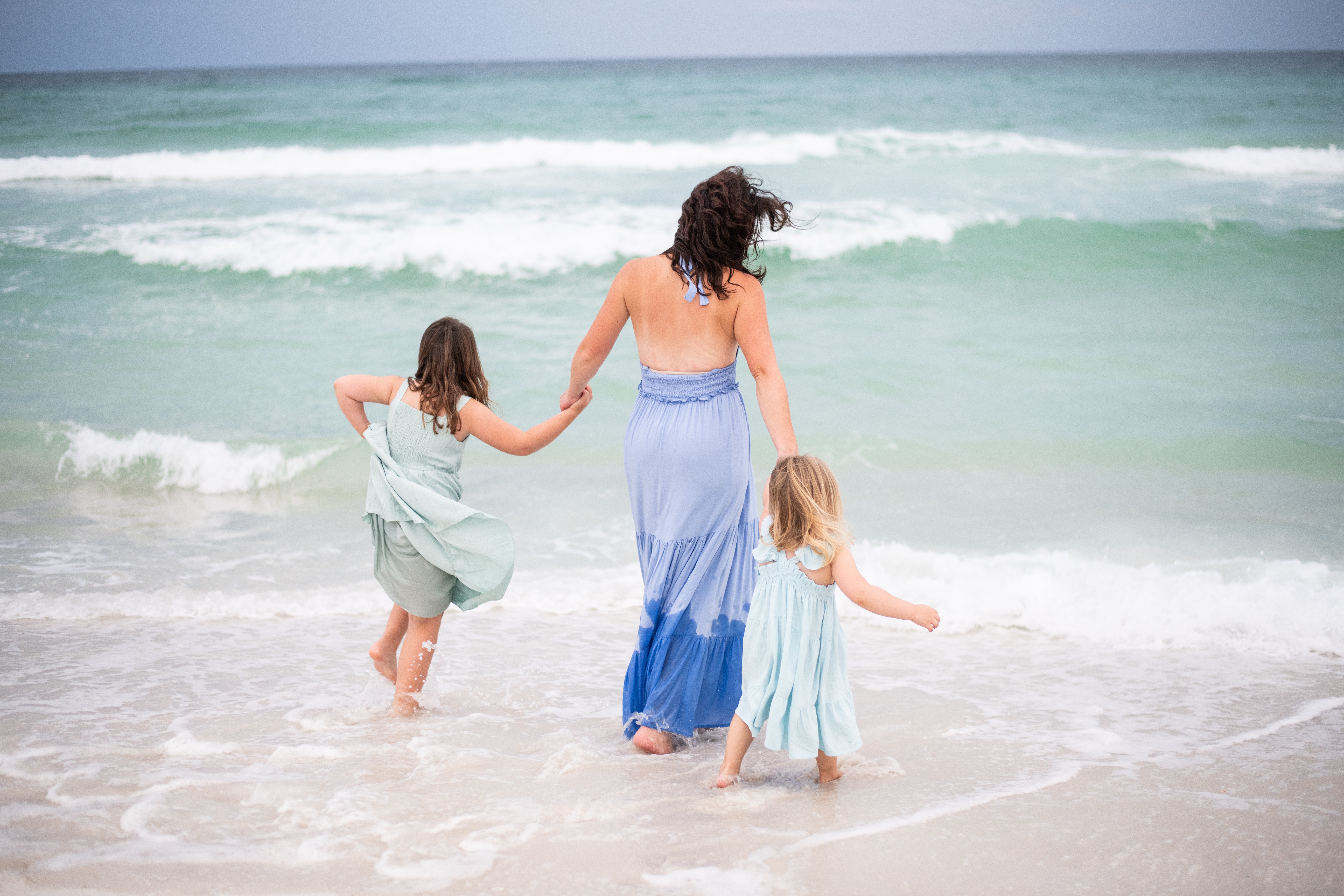 Woman and two children walking into the ocean holding hands at the beach, waves in the background.
